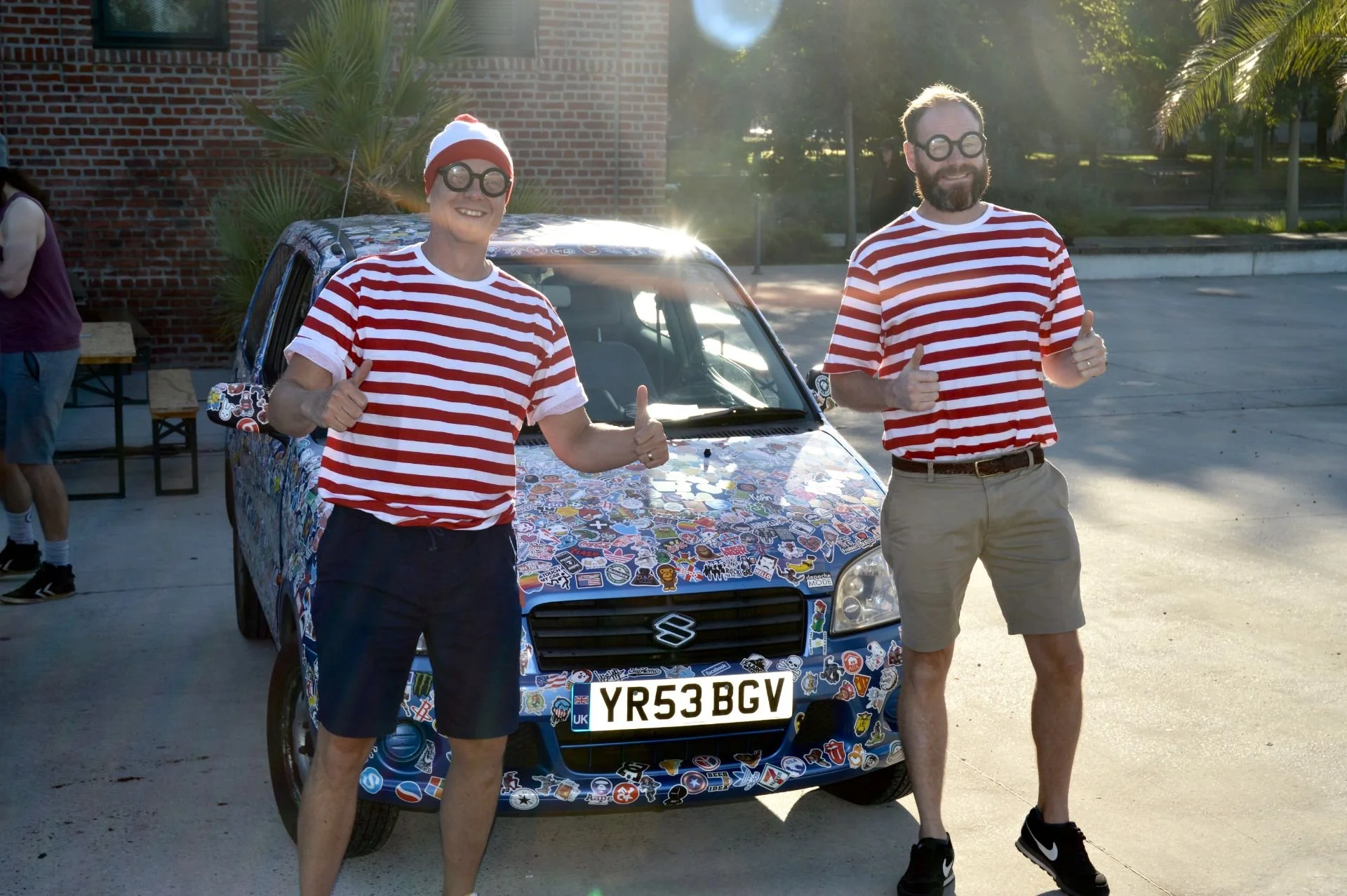 Two men wearing red and white striped shirts and glasses standing in front of a decorated car, showing thumbs up during daytime.