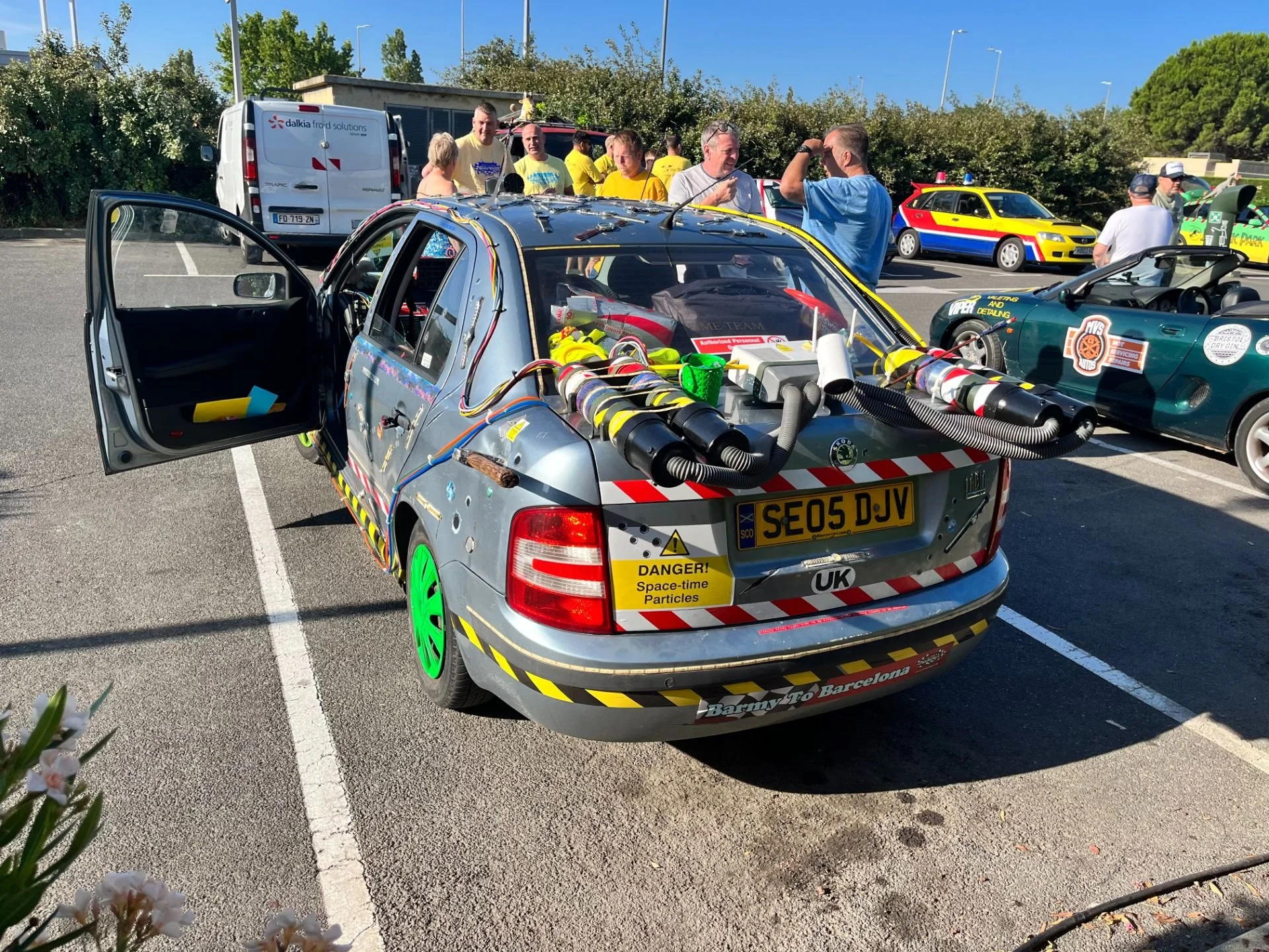 A modified car with various tubes and wires, bright green wheels, and warning stickers, parked in a lot with people gathered around, including a yellow and red vehicle and a dark green convertible nearby.