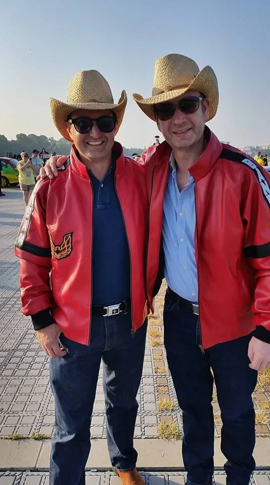Two men in red leather jackets, wearing cowboy hats and sunglasses, standing outdoors with other people and cars in the background.