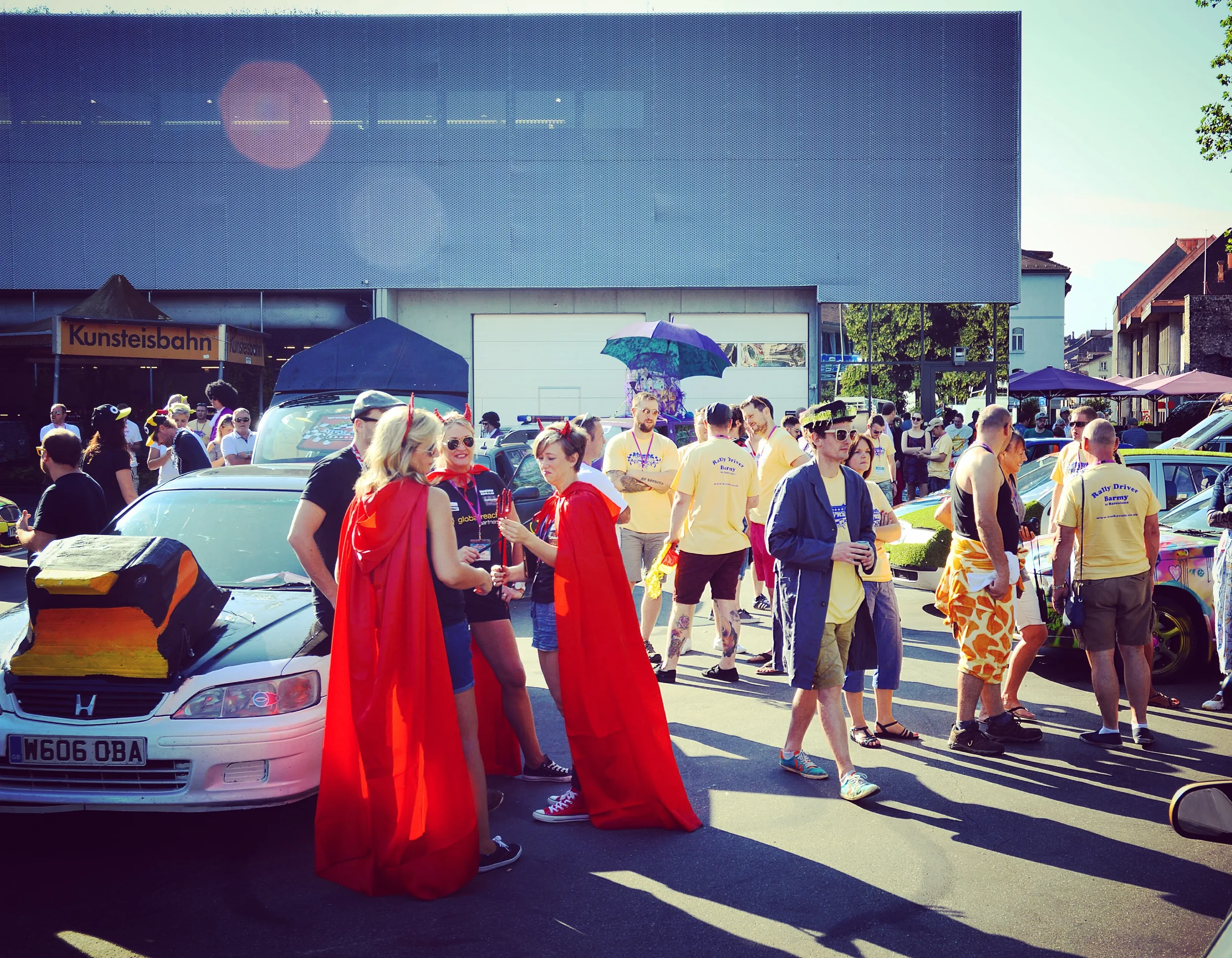 People gathered at an outdoor event or festival on a sunny day, with some dressed in costumes and casual attire, and a car parked in the foreground.