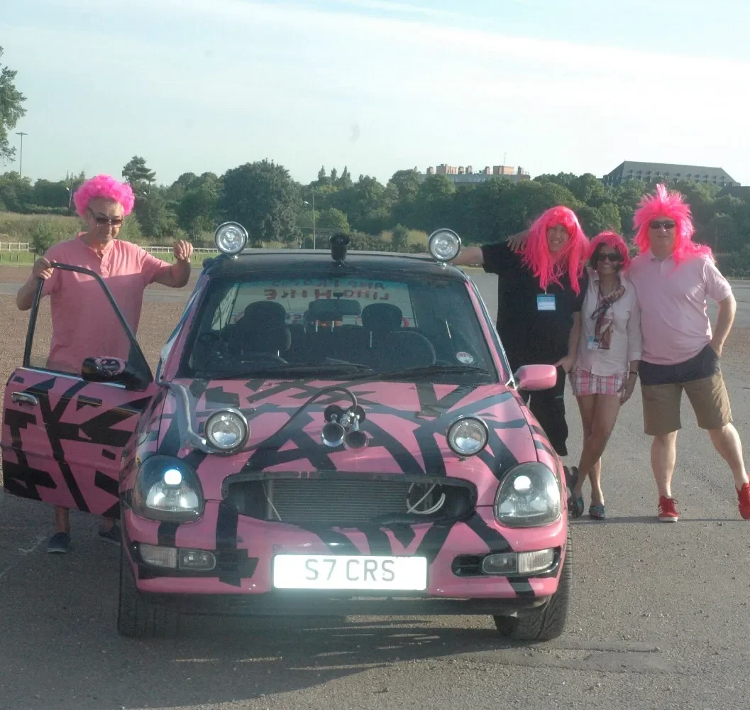 Four people wearing pink wigs and pink clothes posing with a pink and black decorated car in an open area