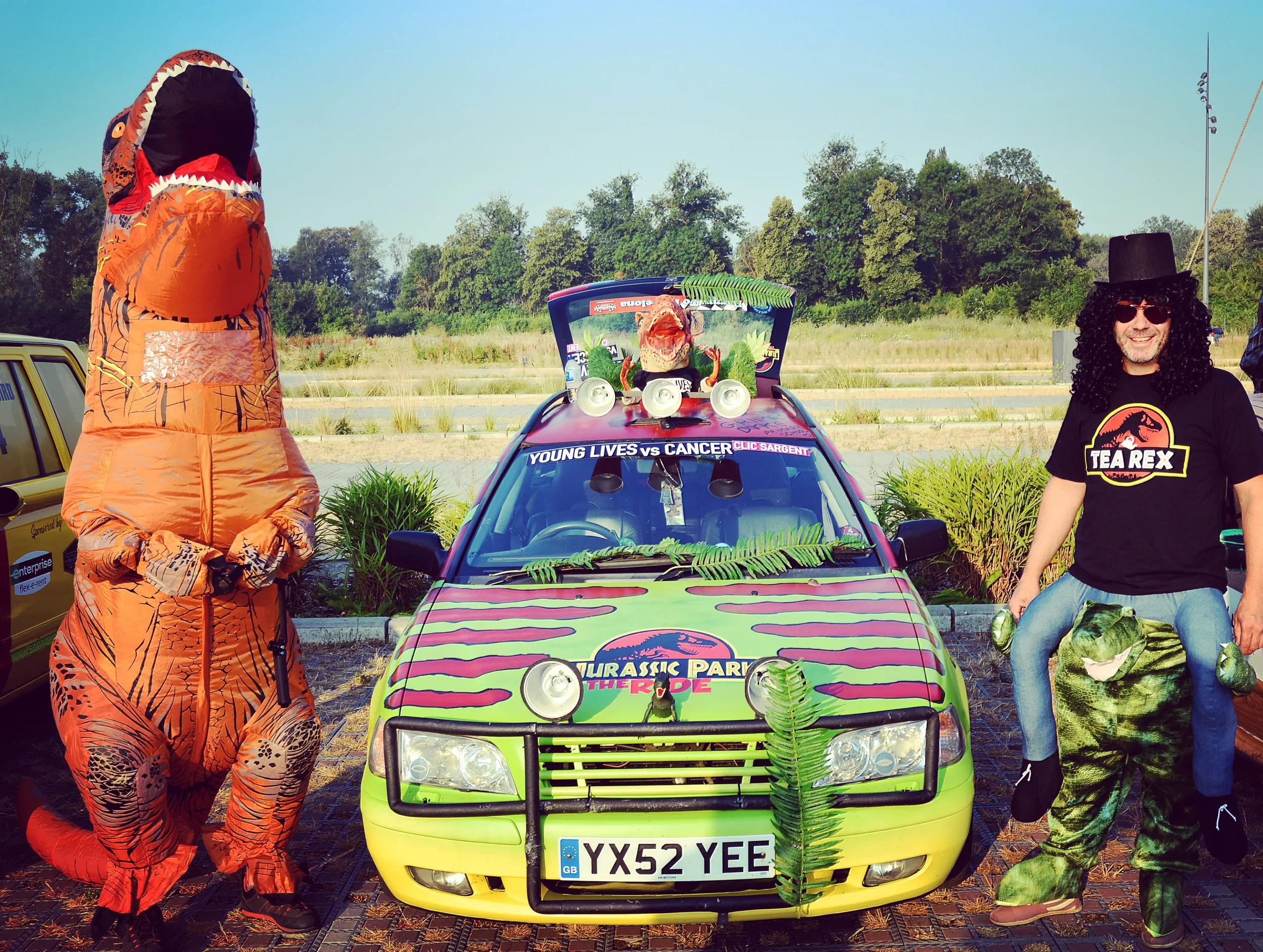 People dressed in dinosaur costumes and a person in sunglasses in front of a decorated vehicle with Jurassic Park theme, set outdoors on a sunny day.