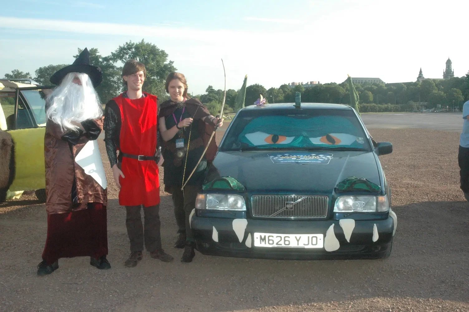 Three people dressed in costumes standing next to a decorated car with large googly eyes and a mouth painted on the front, outdoors on a clear day.