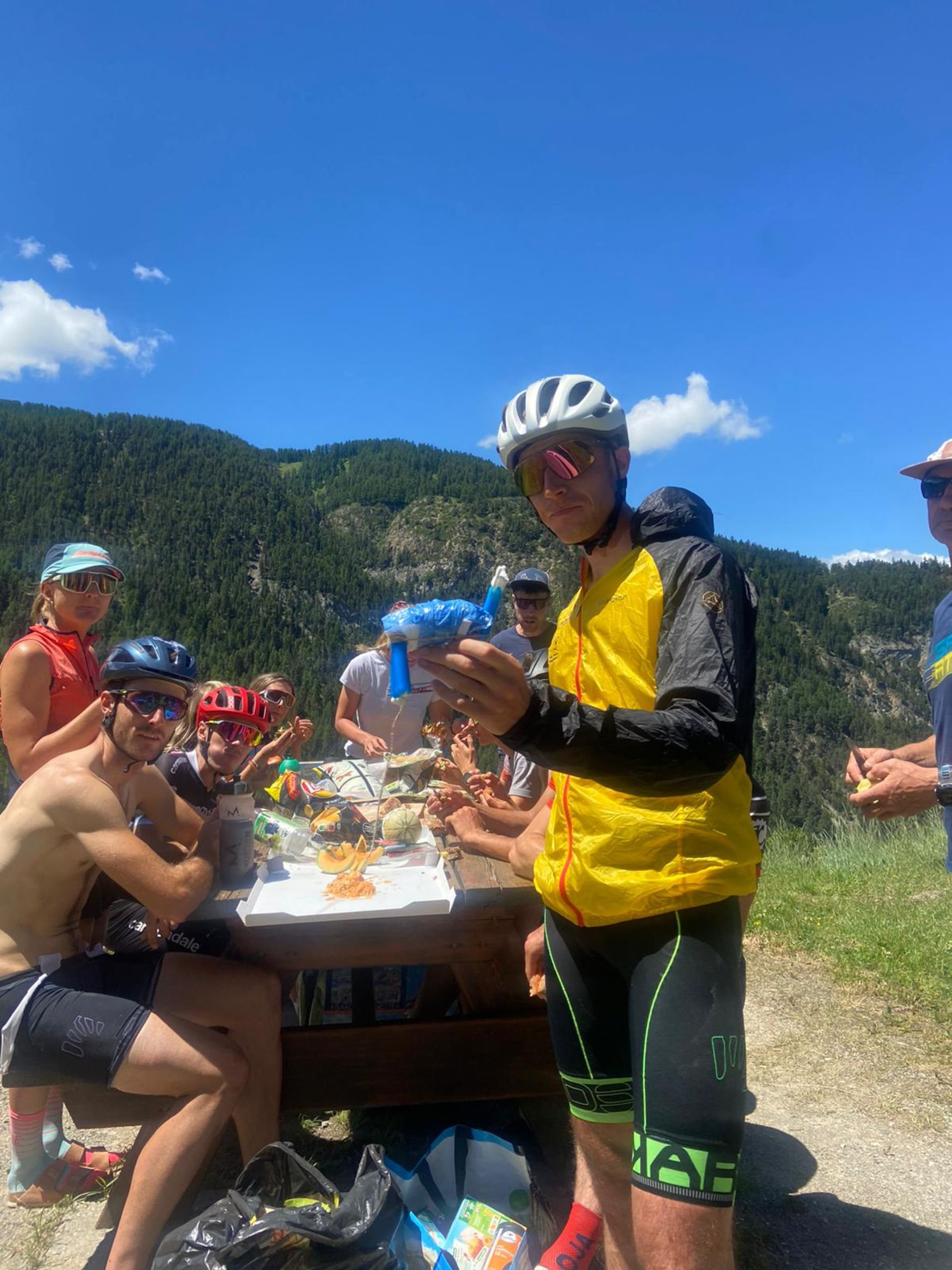 Group of cyclists taking a break at a picnic table outdoors, surrounded by trees and mountains, on a sunny day.
