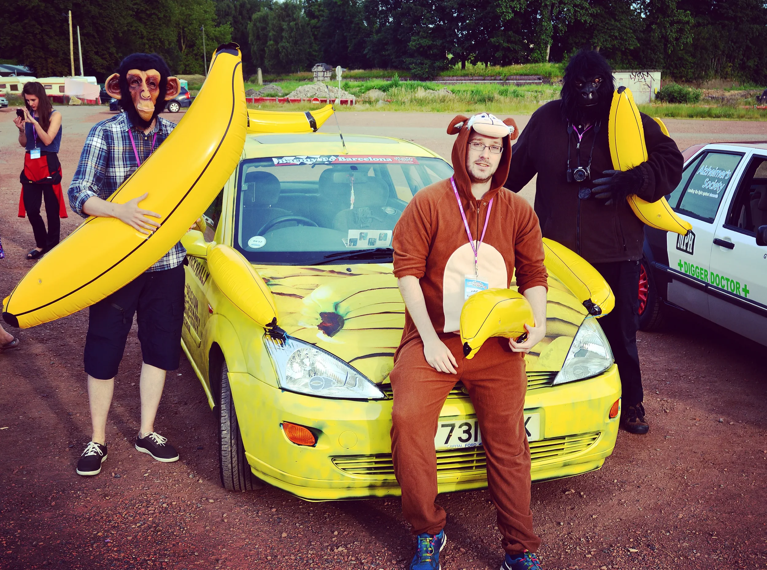 Three people dressed in animal costumes posing with a yellow car decorated with banana-themed accessories. One person is dressed as a monkey, holding a large inflatable banana; another as a bear, holding a smaller inflatable banana; and the third in 