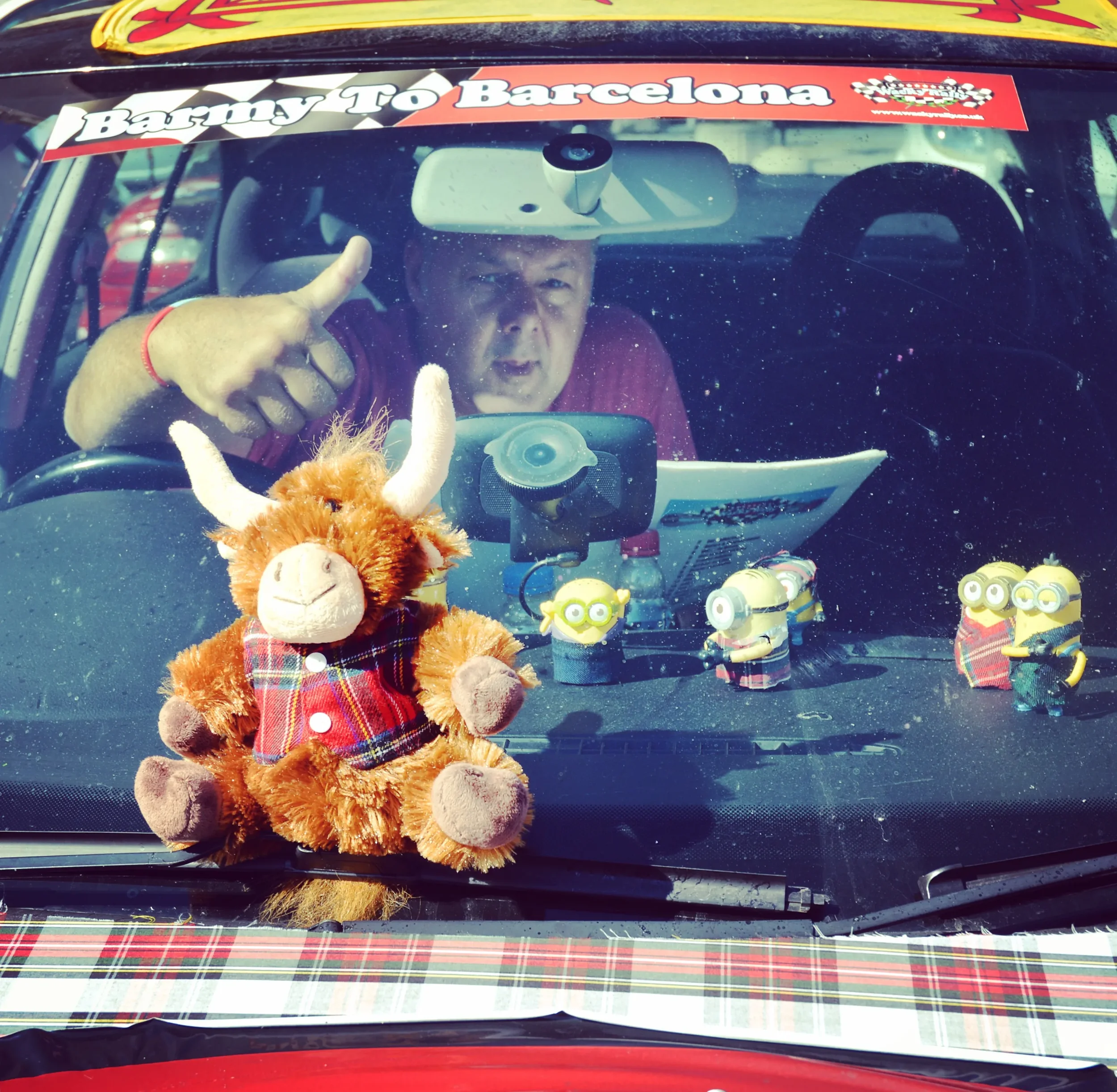 A man inside a car with a sign on the windshield that says 'Barrry To Barcelona.' The top of the car has a tartan pattern, and the dashboard displays plush and minion toys, including a plush teddy bear dressed in a tartan vest and comic character fig