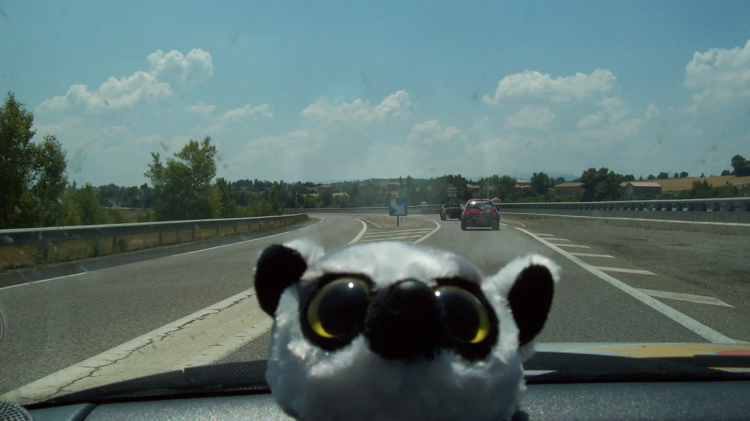 A plush toy with big black eyes and a white face, placed on a car dashboard, with a highway and cars in the background.