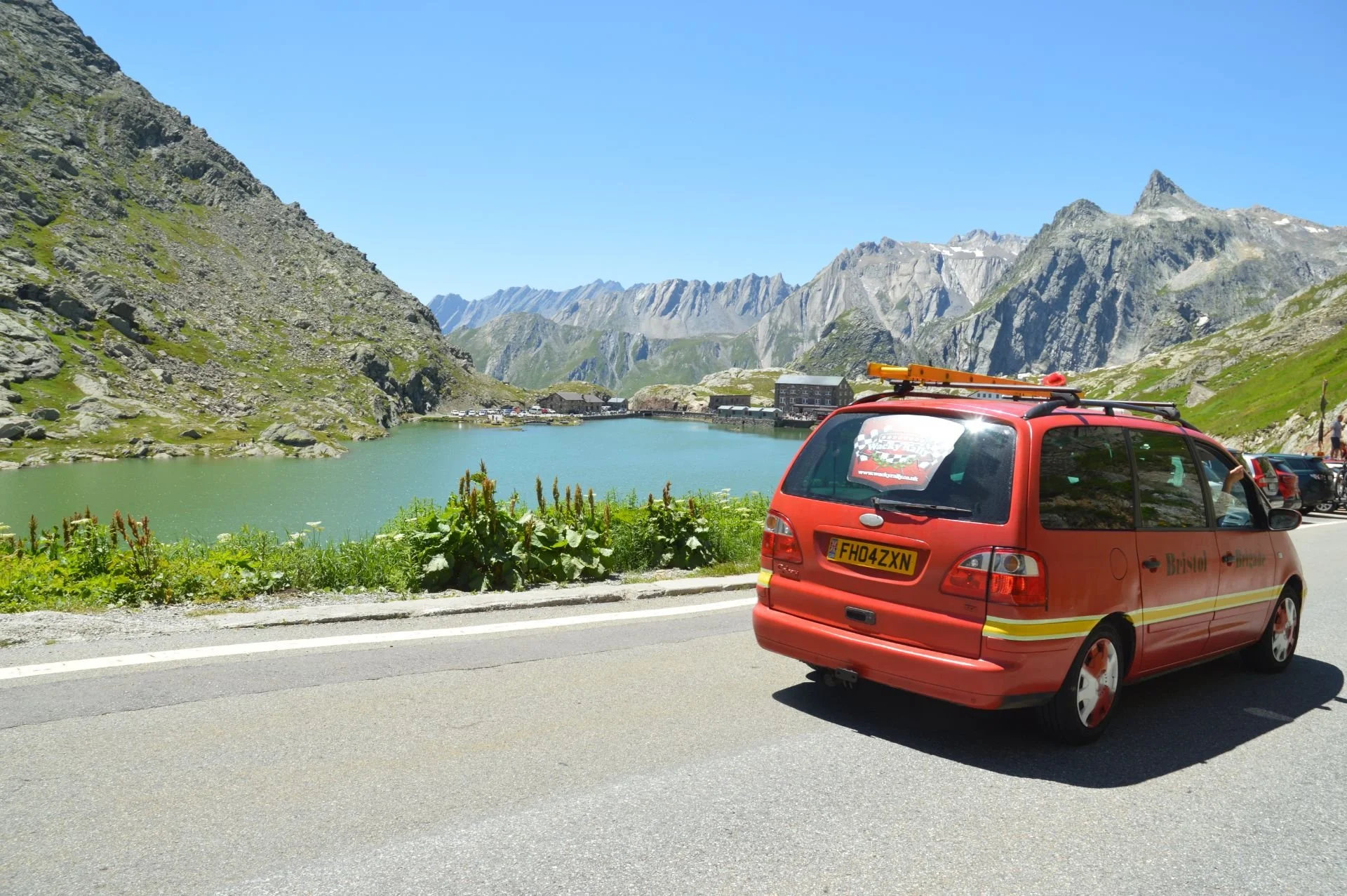 Red emergency vehicle parked on side of mountainous road near a lake, with mountains and a few buildings in the background.
