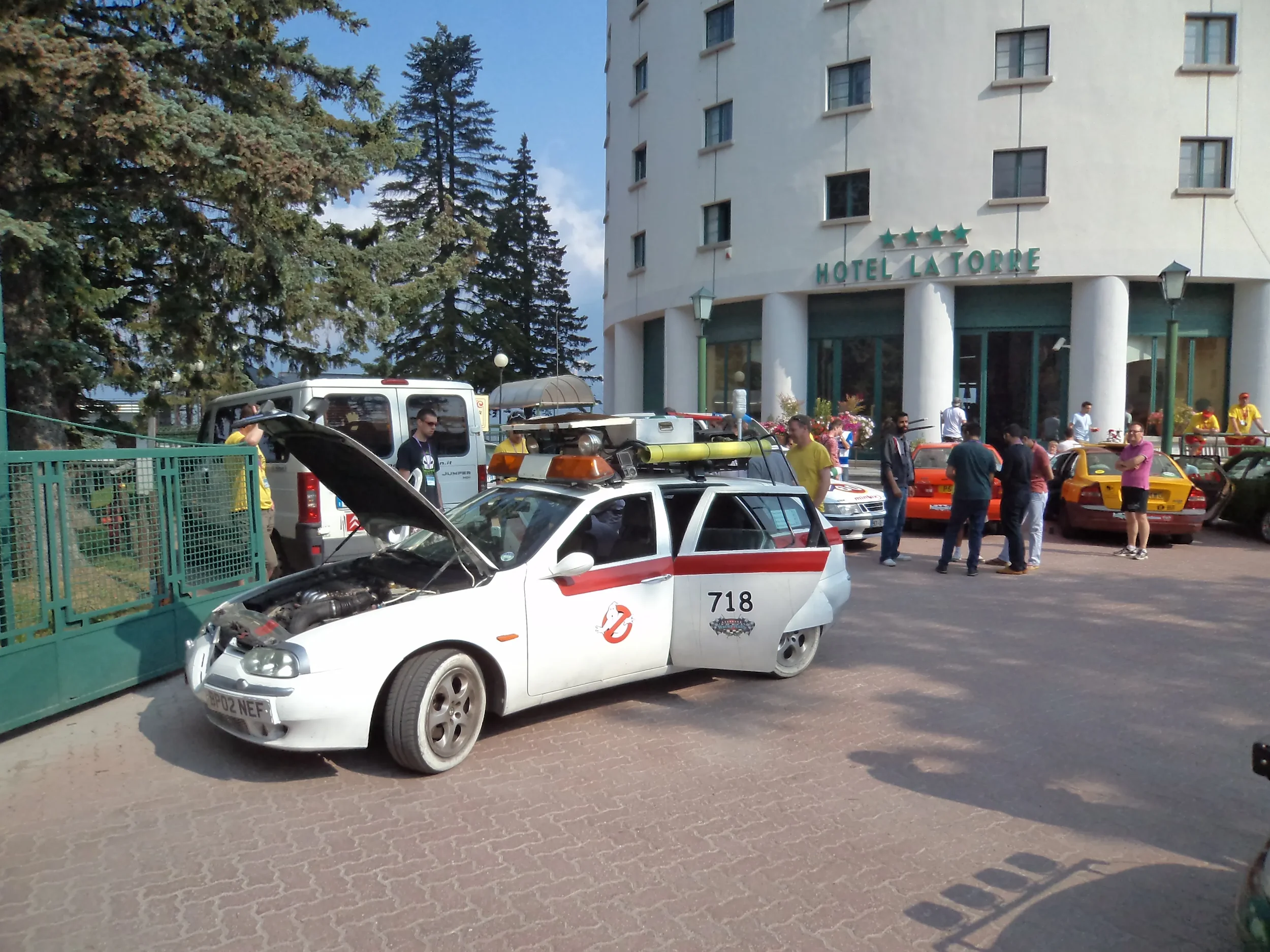 A parody of the Ghostbusters vehicle, modified to resemble the Ecto-1 with Ghostbusters logo, parked outside a hotel with open hood, surrounded by people including some dressed as Ghostbusters, on a sunny day.