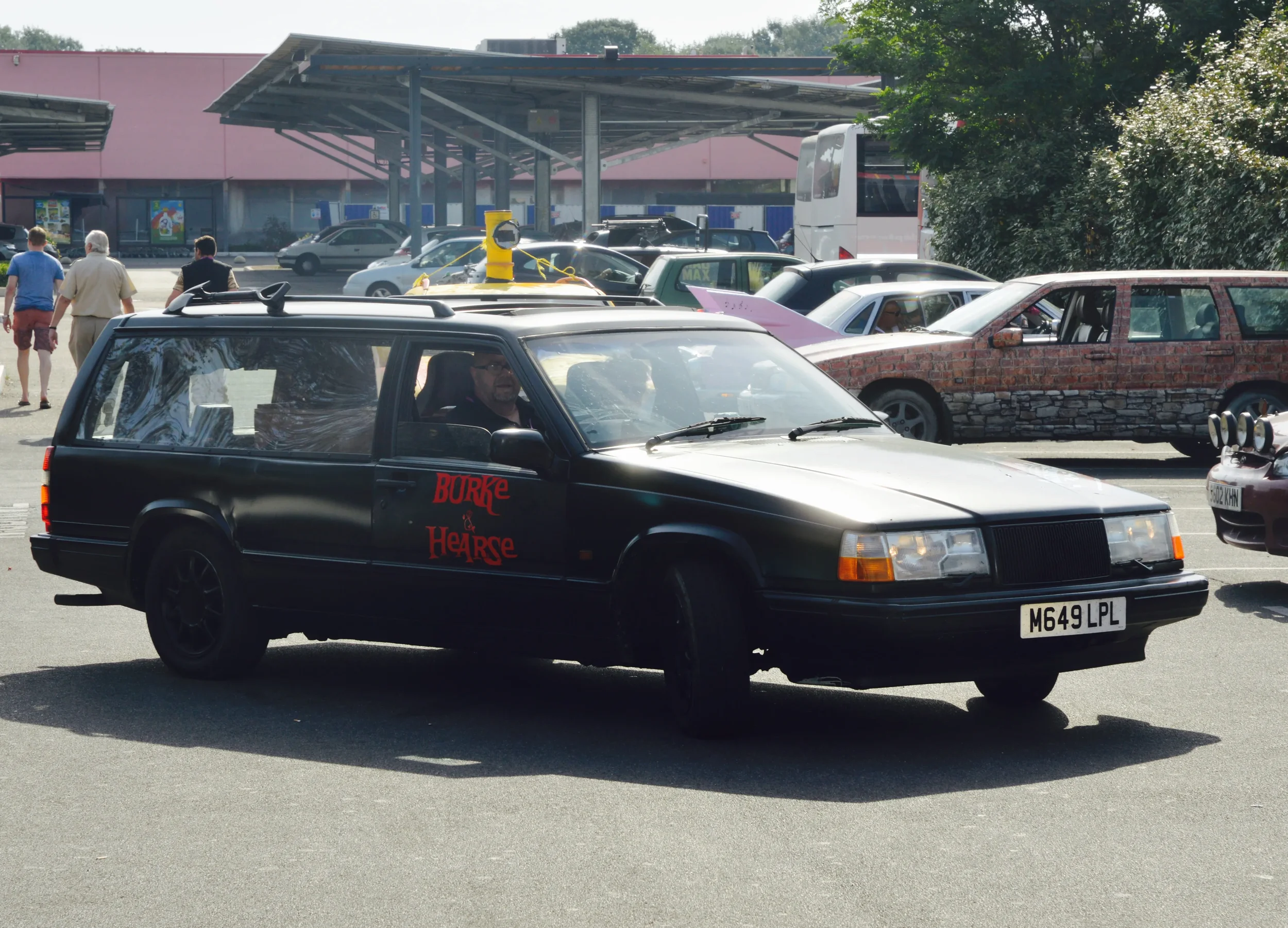 A black station wagon with red text on the side reading 'Burke & Hearse' parked in a lot. The car has a British license plate, and there's a person inside. In the background, other cars are parked, some with creative modifications, and a few people a