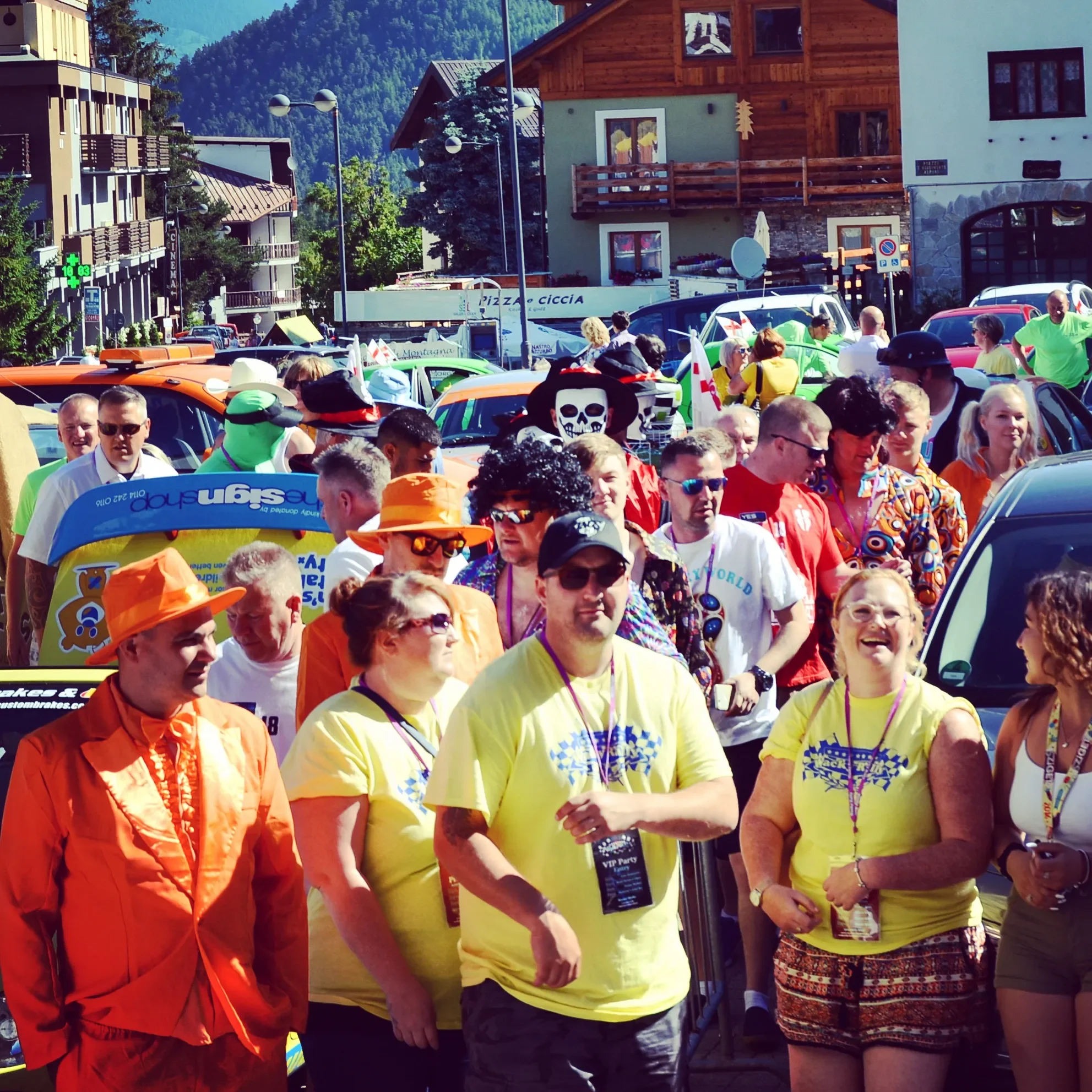 Crowd of people at an outdoor event or festival, many wearing colorful clothing and accessories, some with costumes like skeleton and clown face paint, hats, and sunglasses, with mountains in the background.