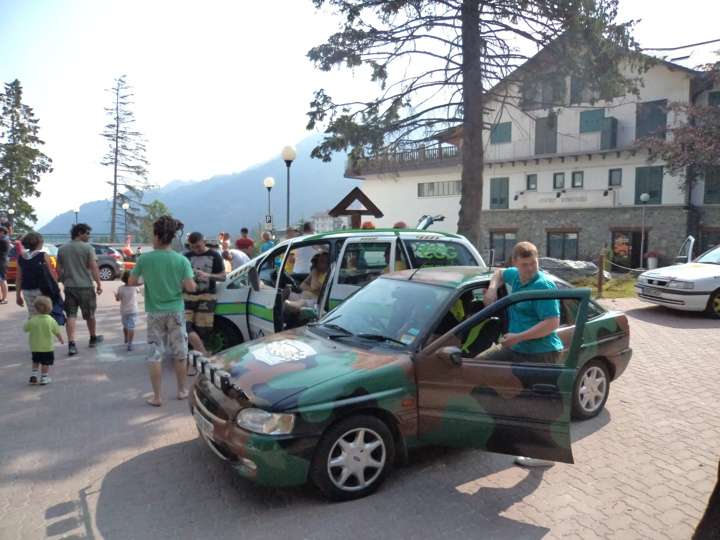 People gathered around cars, including one with a camouflage paint job, in an outdoor parking area near a building with a mountain backdrop.