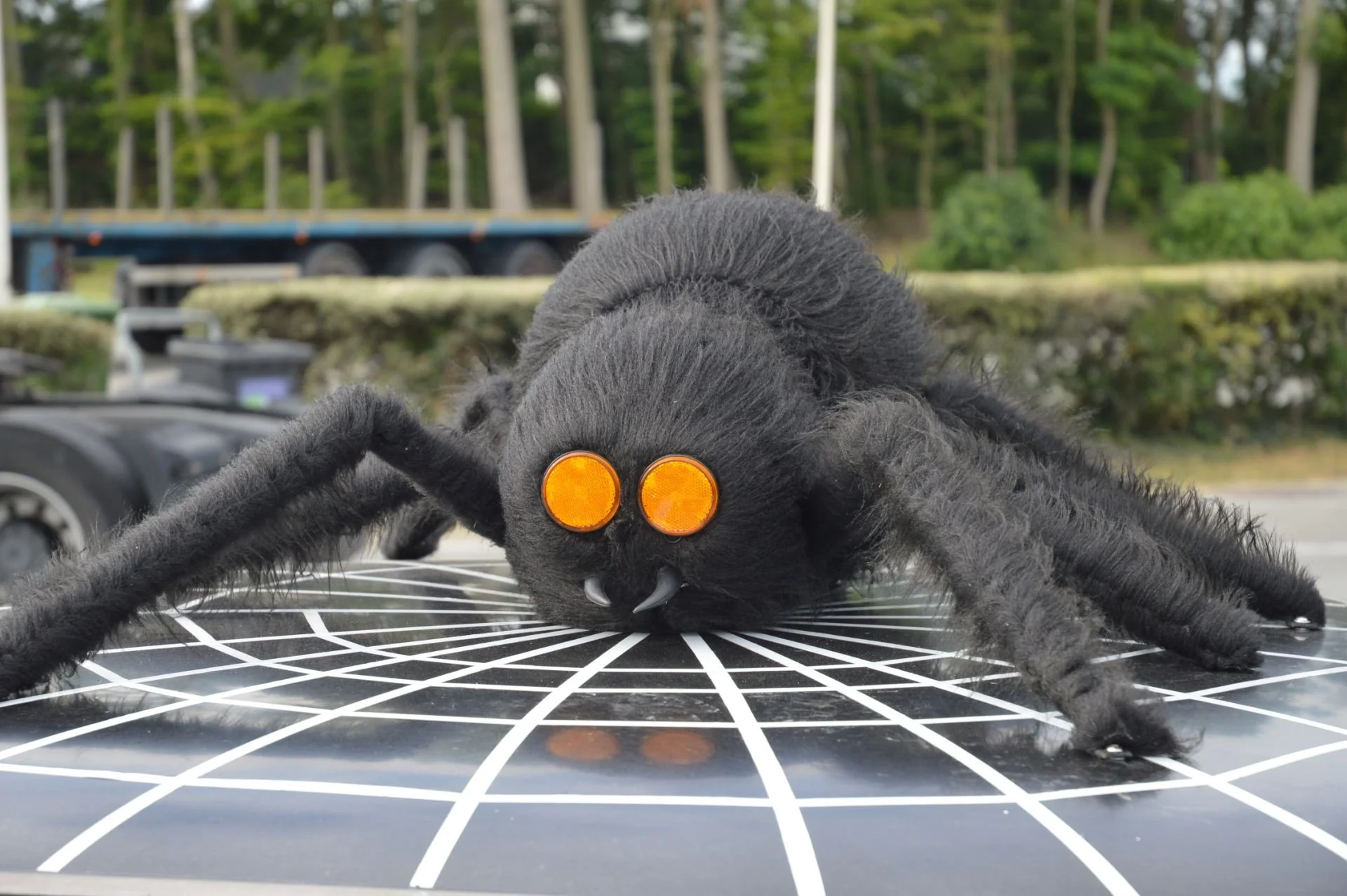A large, black, spider-shaped plush toy with orange eyes and fangs, lying on a black and white grid-patterned surface outdoors with trees and vehicles in the background.