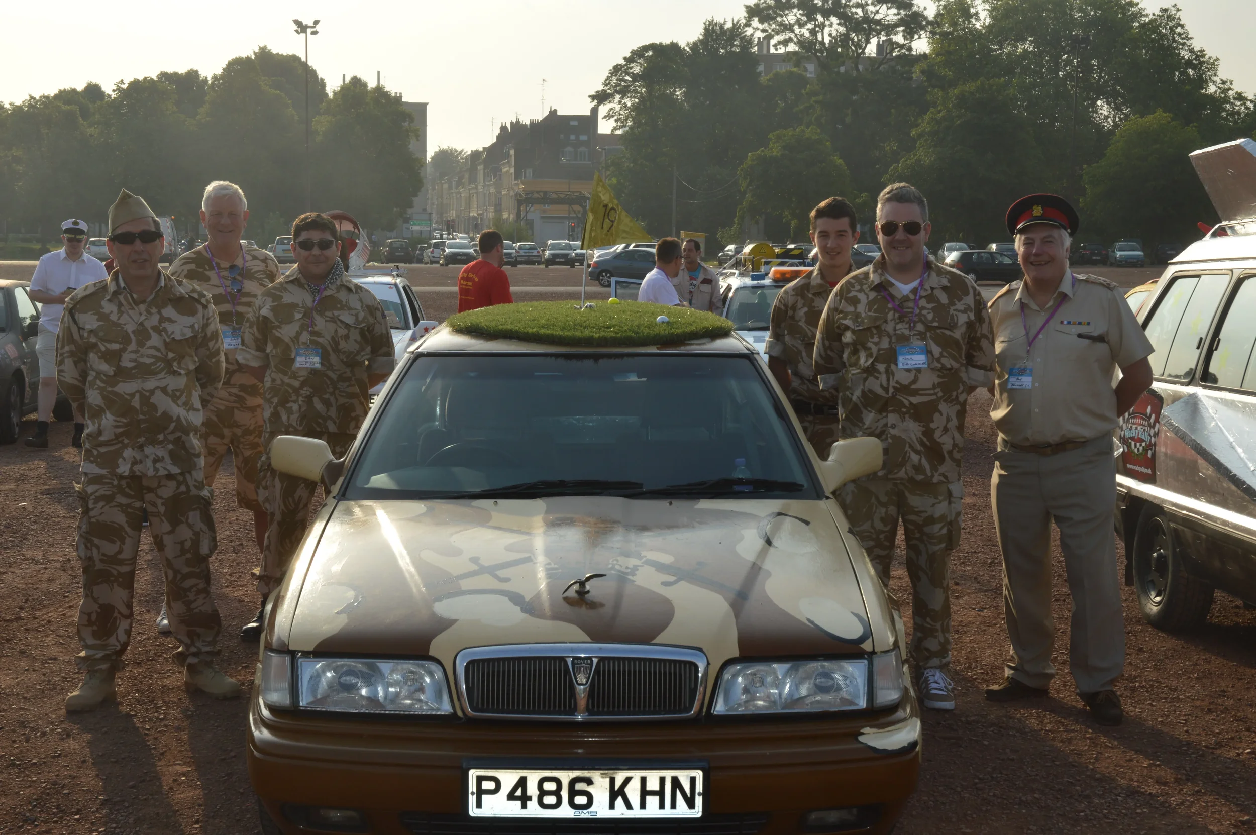 Group of military personnel in camouflage uniforms and a man in a light-colored uniform standing beside a camouflage-painted car with a grass-like canopy and a yellow flag, surrounded by other vehicles in an outdoor setting.