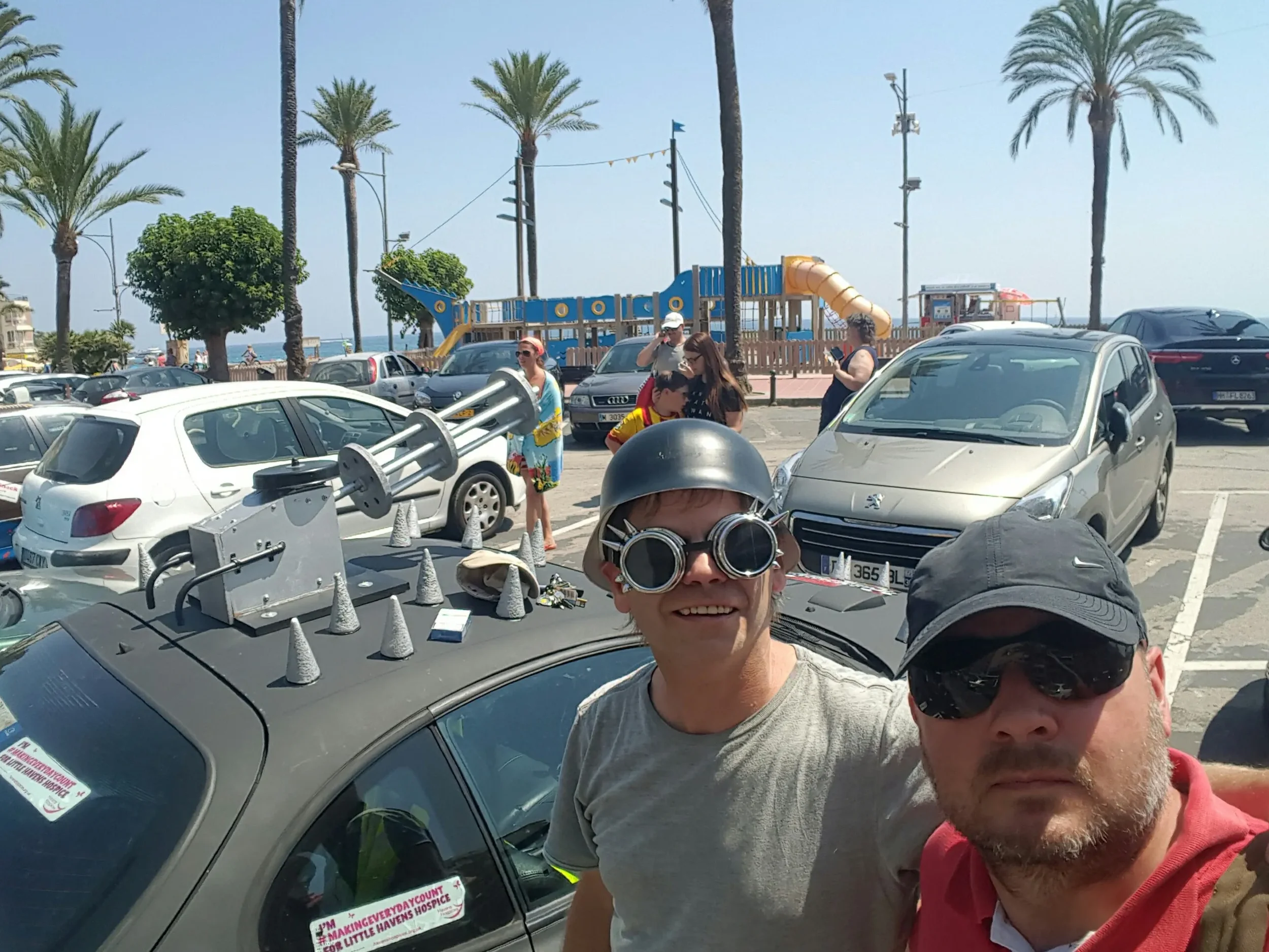Two men taking a selfie next to a decorated car with plastic cones on the roof, people and parked cars in the background near a beach with palm trees and a playground.