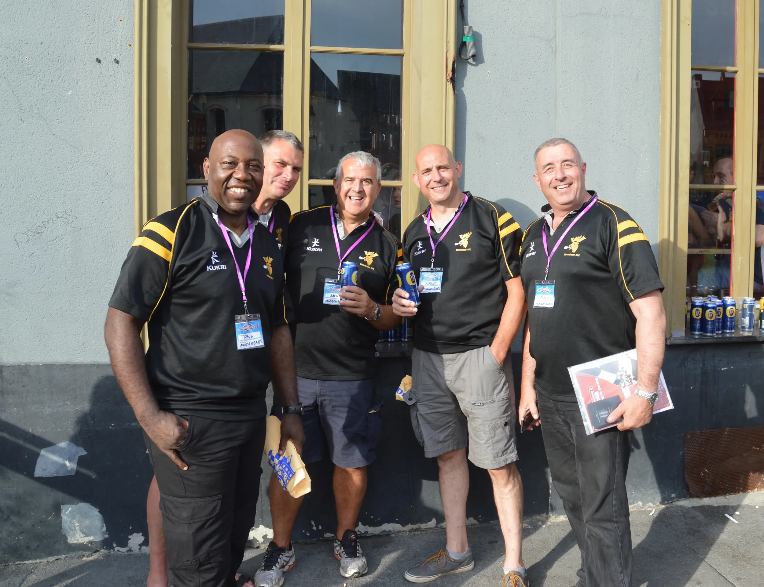 Group of five men standing outdoors, wearing black sports jerseys with yellow accents, smiling at the camera, holding drinks, and wearing conference badges around their necks.