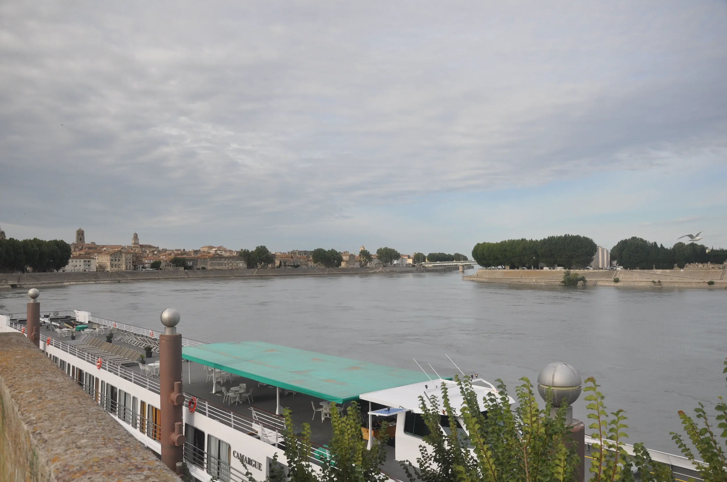View of a river with a cityscape in the background, a docked boat with a green canopy in the foreground, and trees along the riverbank under a partly cloudy sky.