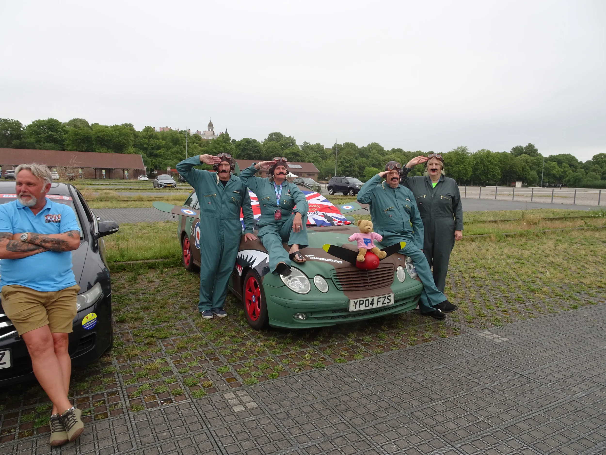 Four people dressed as pilots in blue jumpsuits saluting beside a green car decorated with a teddy bear and a large plush toy, with a man standing to the left in a blue shirt with tattoos, in an outdoor parking lot on a cloudy day.