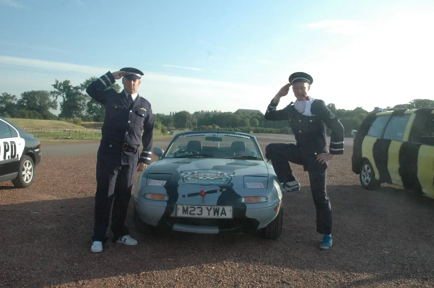 Two men dressed in police costumes salute in front of a small race car, with police cars visible on the sides, in an outdoor setting during daytime.