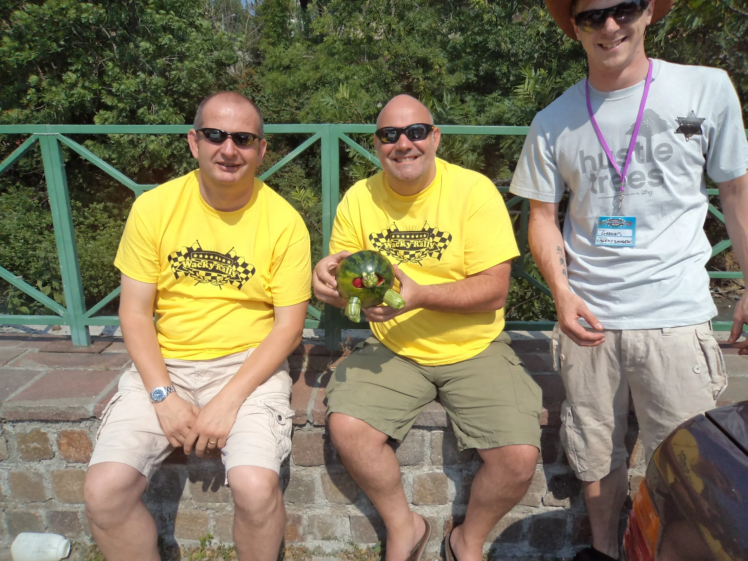 Three men sitting on a brick wall outdoors, wearing sunglasses and casual clothing. The man in the middle is holding a green turtle-shaped object with a red nose. The man on the right is wearing a gray T-shirt with a name tag around his neck.