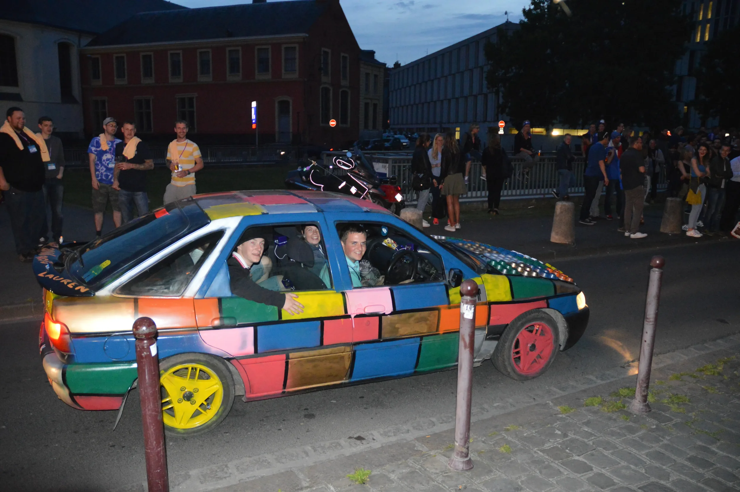 A brightly painted car with a multicolored, geometric pattern on its body and red wheel rims is parked on a city street at dusk, with three smiling people inside, and a crowd of people standing along the sidewalk in the background.