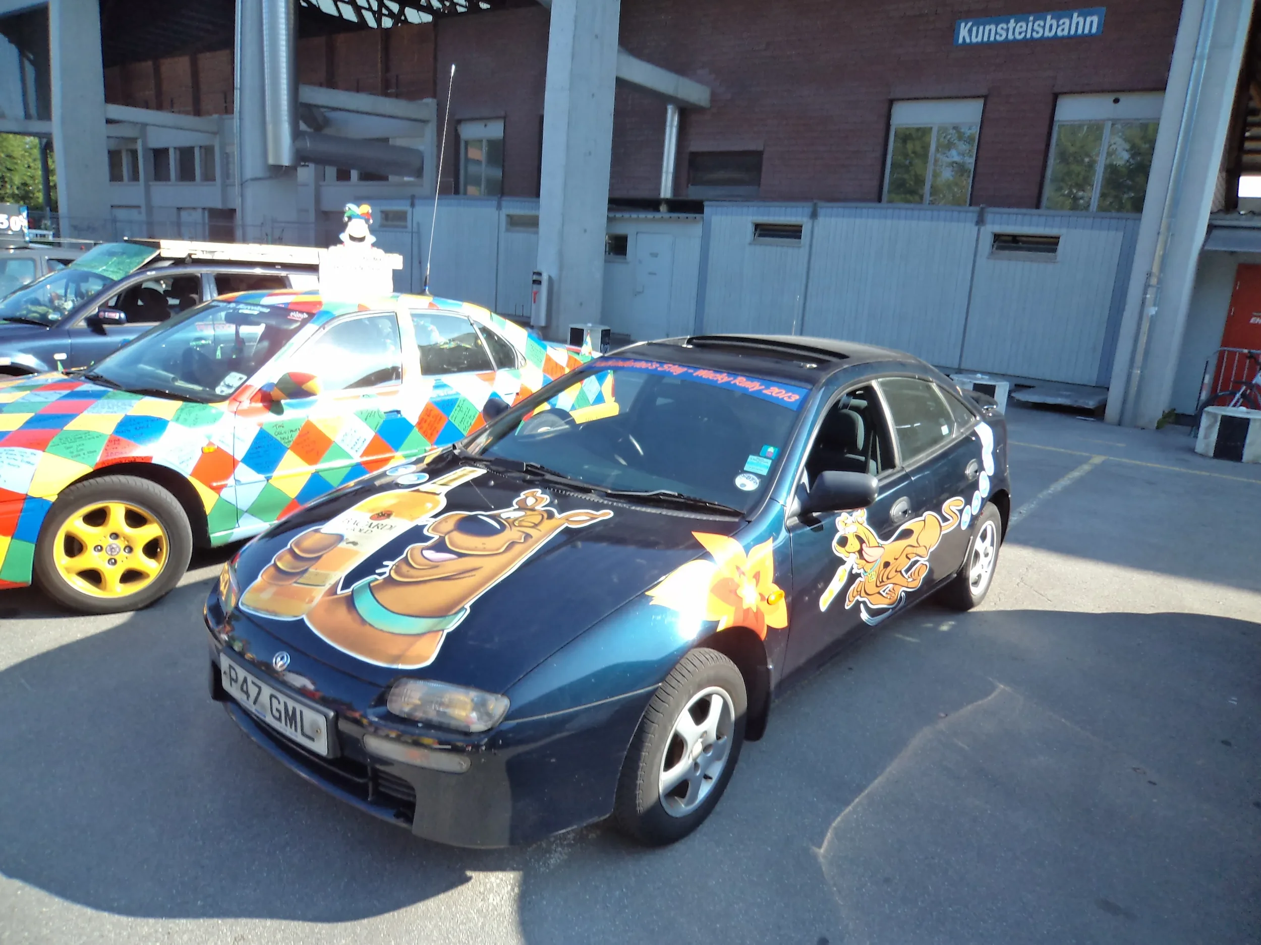 A black car decorated with cartoon characters, including a bear and a dog, parked in an outdoor parking lot next to a colorful sports car with a harlequin pattern. The cars are themed for a promotional or festive event, with banners and signs nearby.