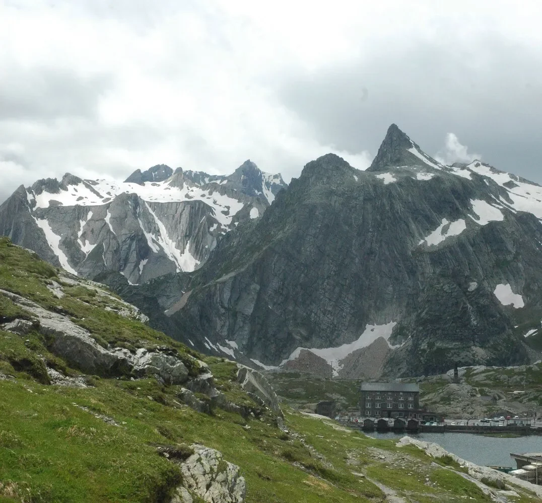 Snow-capped mountains behind a green grassy hillside with rocks, near a body of water with a building and boats at the shore.