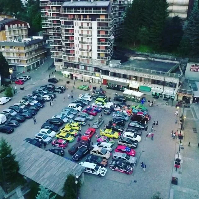 A parking lot in an urban area with numerous colorful cars, surrounded by tall buildings and some people walking around.