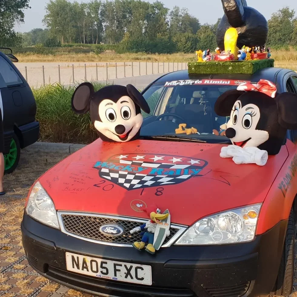 A red Ford car decorated with large Minnie Mouse and Mickey Mouse head mascots on the hood, a giant black swan figure on the roof, and cartoon character Pluto sitting on the front grille, with a racing-themed rally sticker and various signatures on t