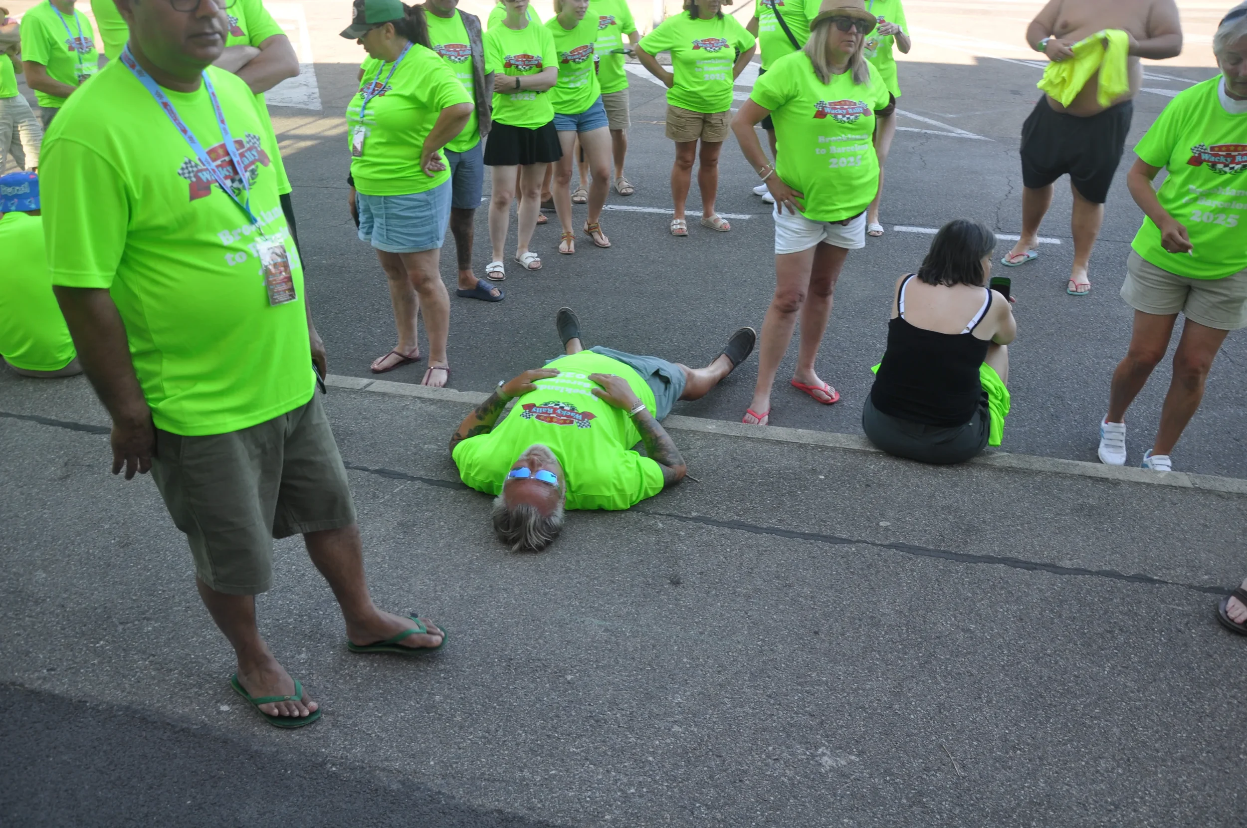 Person lying on the pavement wearing bright green T-shirt, surrounded by a group of people, many also in bright green T-shirts, at an outdoor event or gathering.