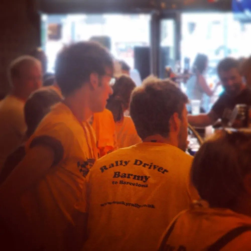 Group of people wearing yellow shirts with the text "Rally Driver Harry to Svalbard" printed on the back, sitting around a table in a dimly lit restaurant or bar.