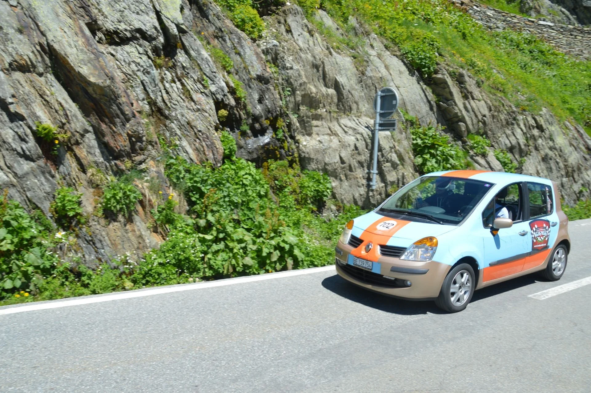 A small rally car driving on a mountain road with rocky and green terrain.