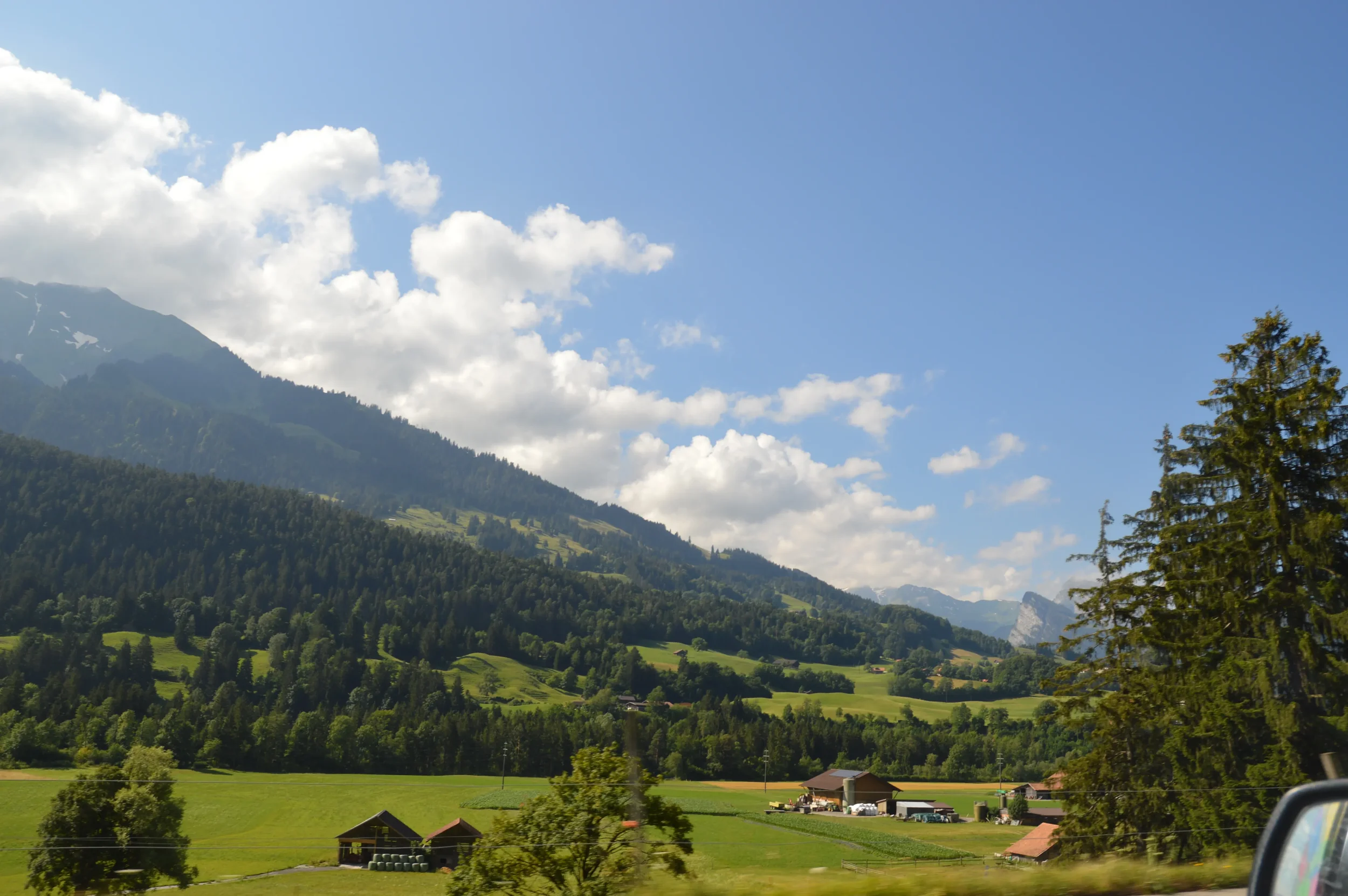 Scenic view of a lush green valley with scattered farm buildings, surrounded by forested hills and mountains under a partly cloudy sky.