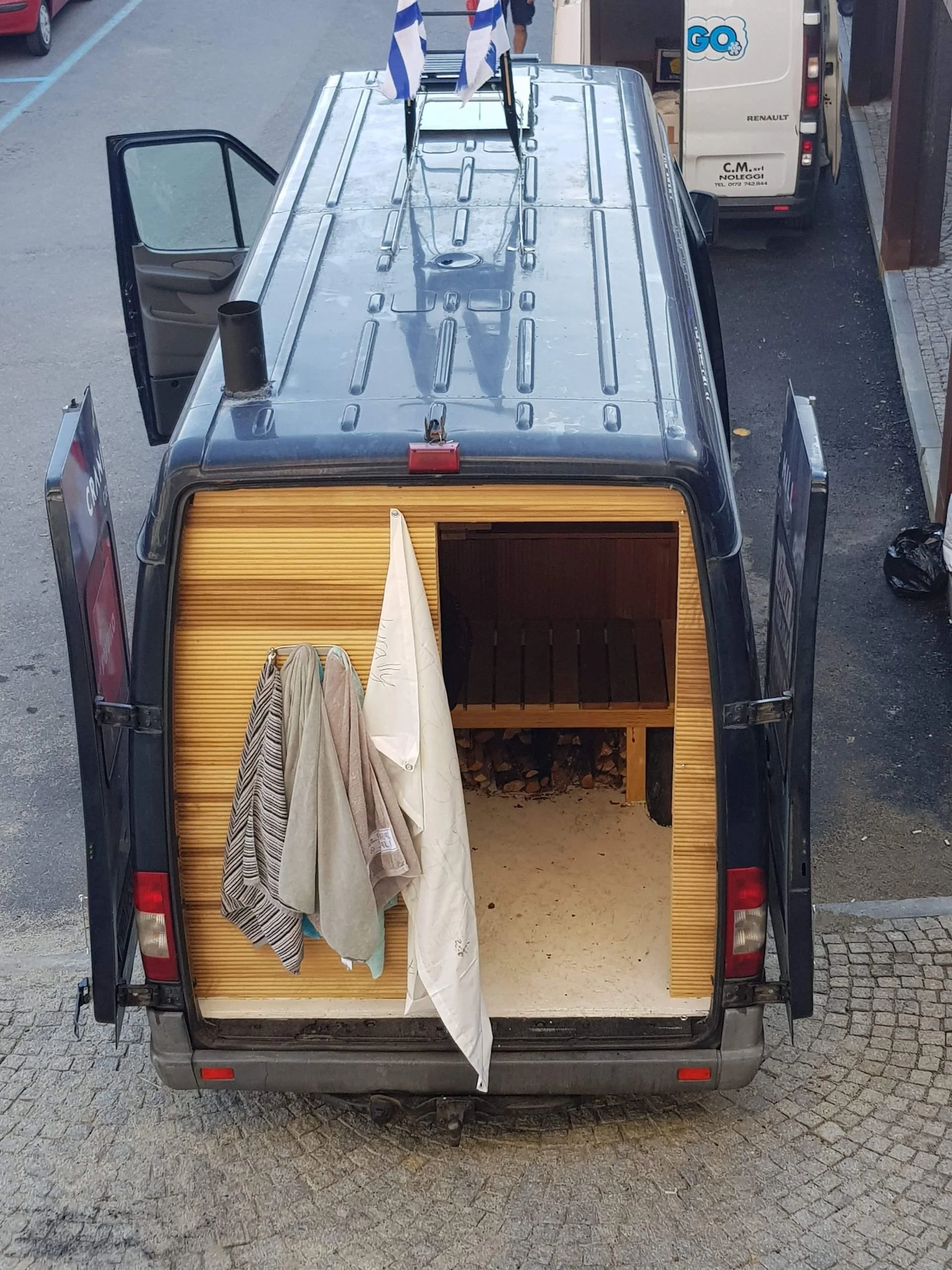 A black van with open rear doors revealing a small sauna interior with wooden benches and a wood storage compartment. Clothes hang on the left door.