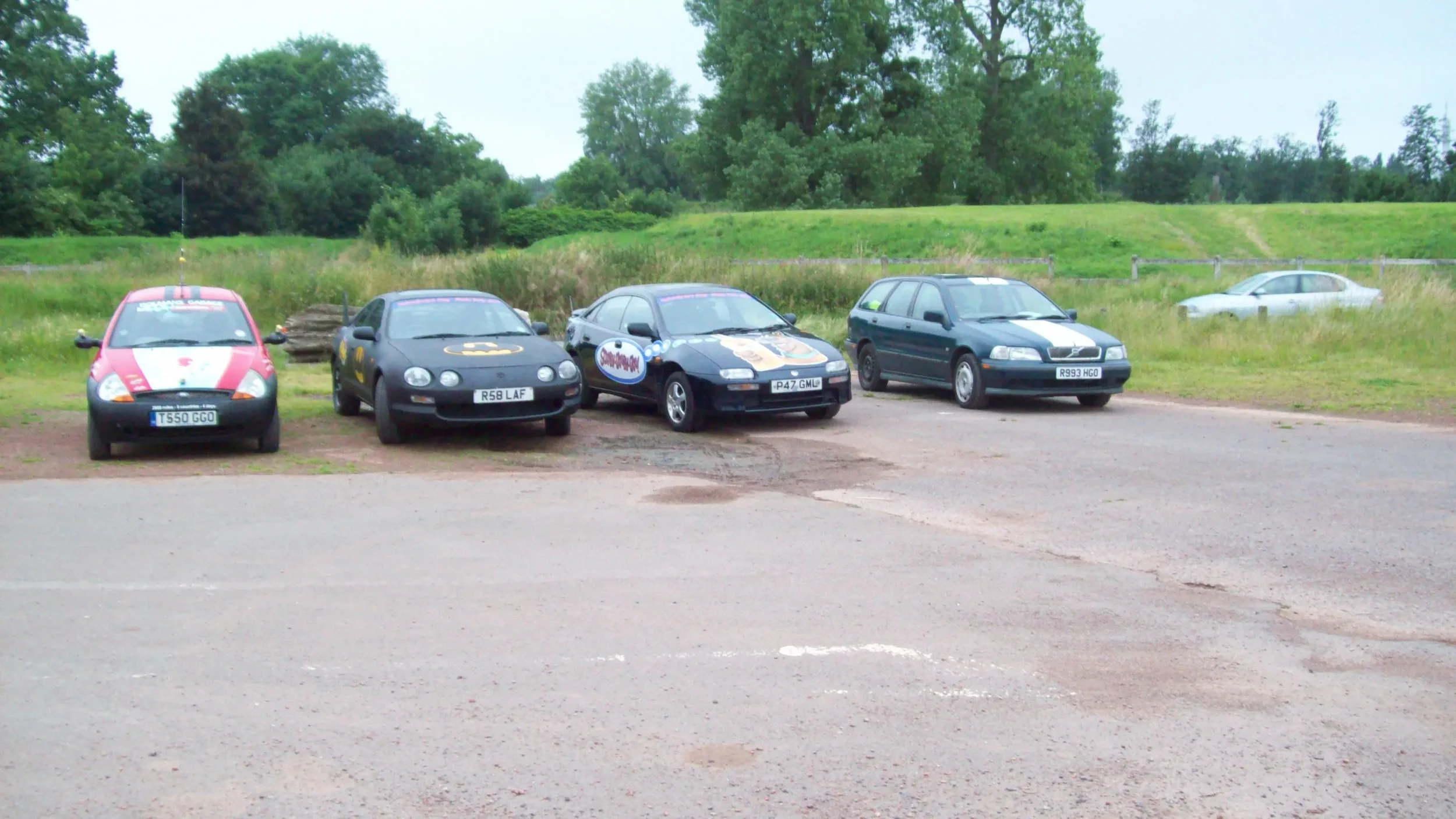Five cars parked in a grassy field with a green wooded area in the background.