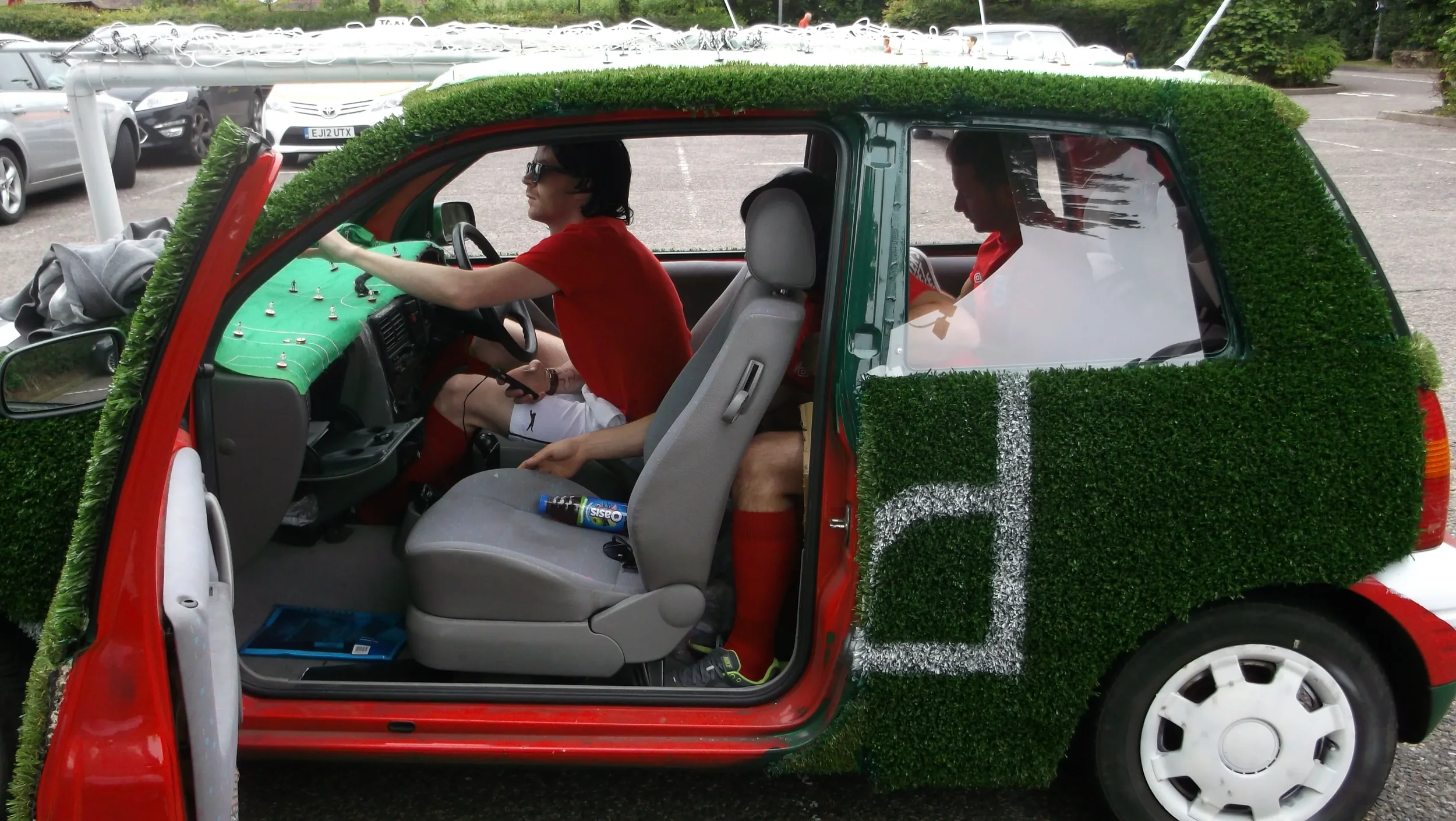 A small car decorated with artificial grass on the exterior and dashboard, parked in a lot with other vehicles. Inside the car, two men are sitting, one in the driver's seat and the other in the passenger seat. The driver is wearing sunglasses and a 