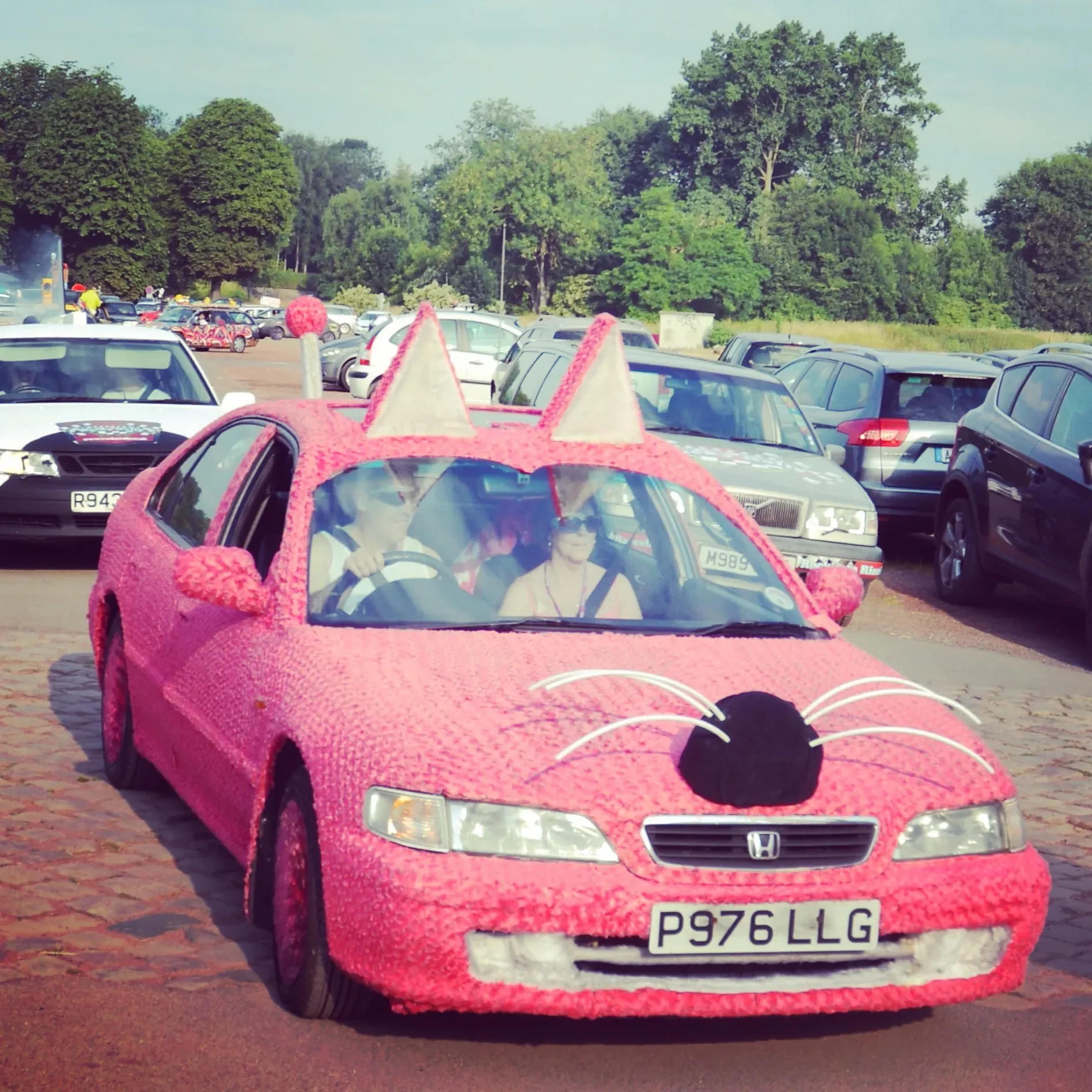 Pink car decorated to look like a cat, with cat ears on the roof, whiskers, and a nose on the hood, parked in a parking lot.