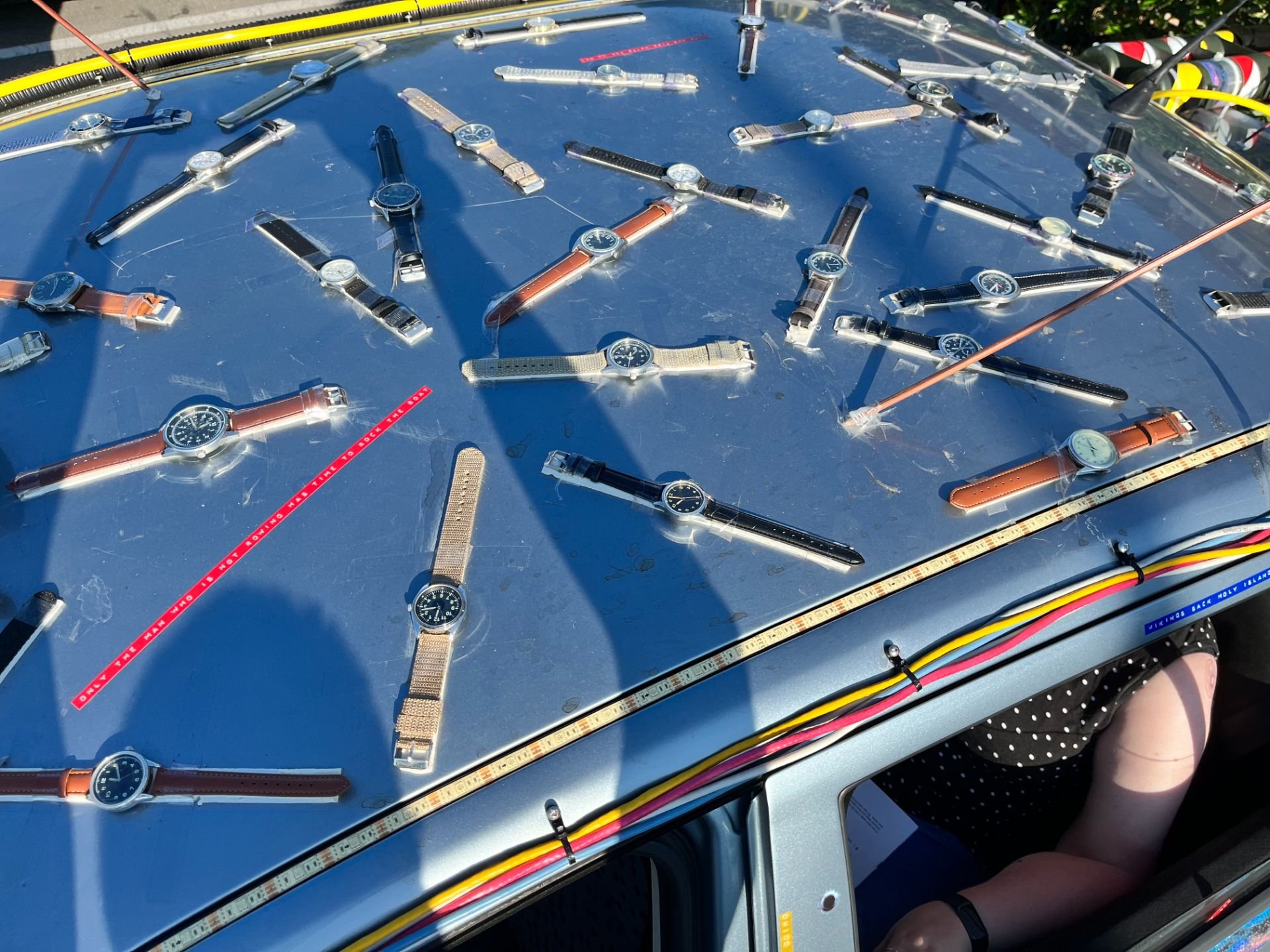 Multiple wristwatches placed on a blue metallic surface, some with metal or leather bands, in sunlight at an outdoor event.