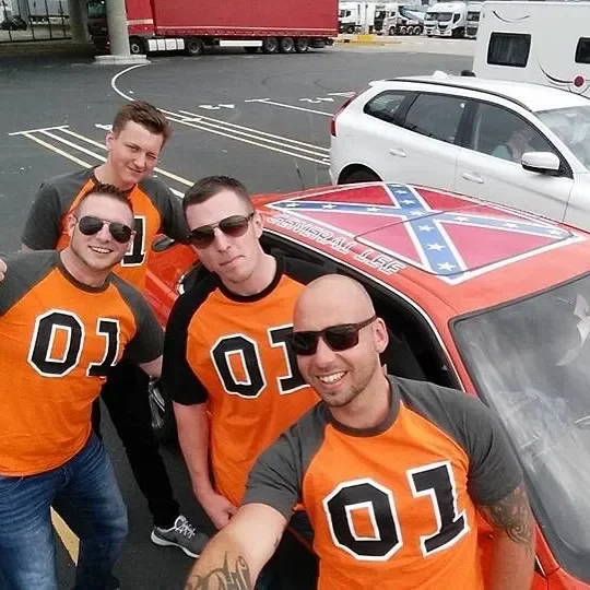 Four men wearing matching orange and black sports jerseys with the number 01 on the front, taking a selfie outside in a parking lot next to a car with a Confederate flag and other markings on the hood.