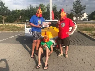 Three people wearing chicken masks, with two standing and one sitting, posing in a parking lot with a green train ride vehicle.