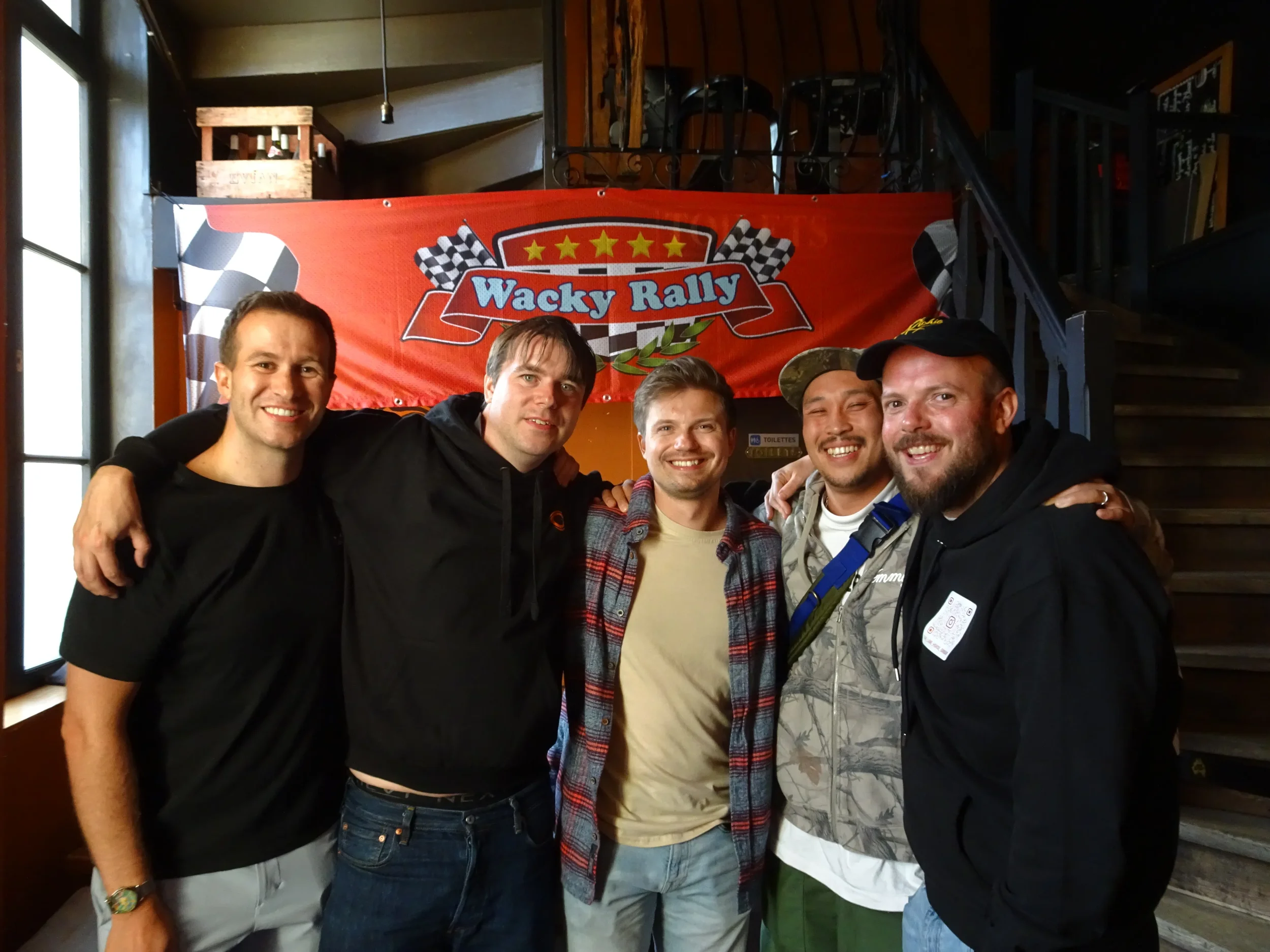 Five men standing together with their arms around each other, smiling in front of a red banner that reads 'Wacky Rally' with checkered flags and stars, inside a room with dark wood stairs and a window.