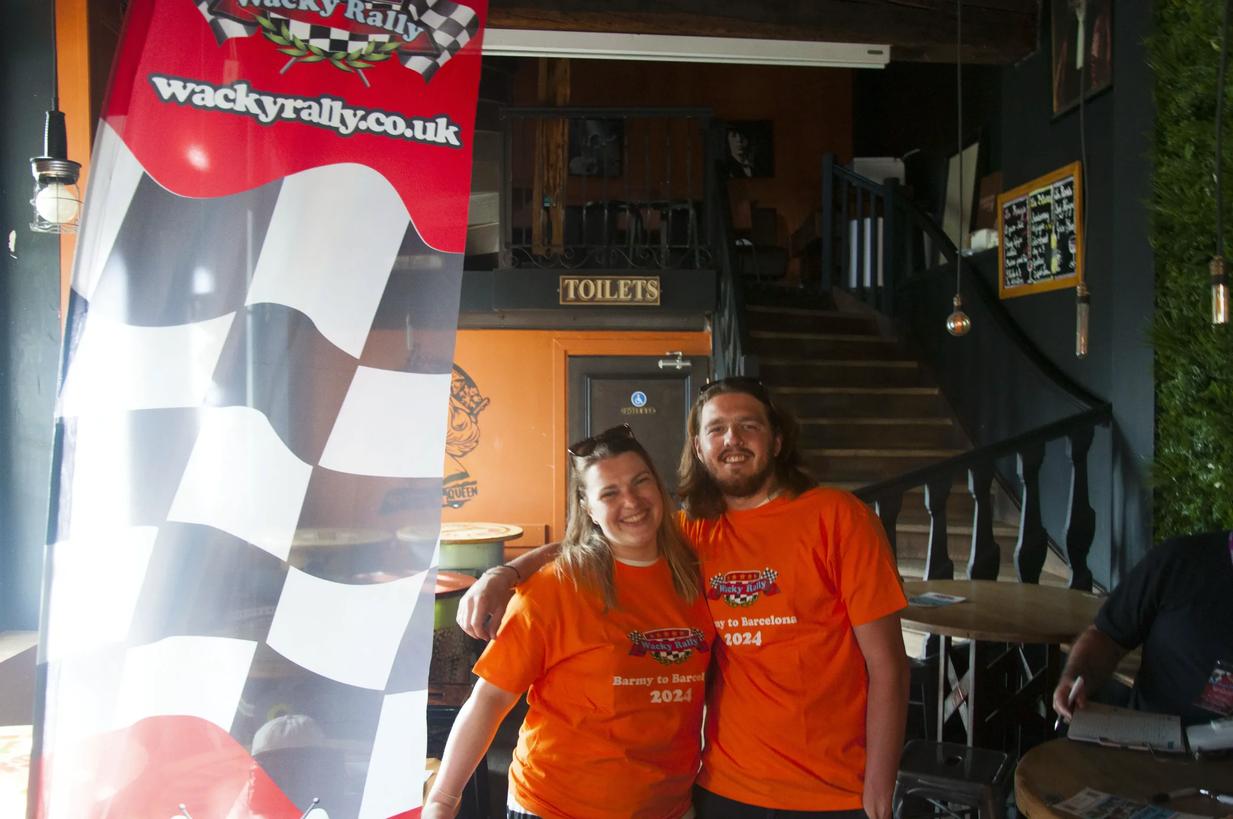 Two smiling people, a woman and a man, standing together wearing matching orange T-shirts, inside a rally-themed venue with a checkered flag banner and a staircase in the background.