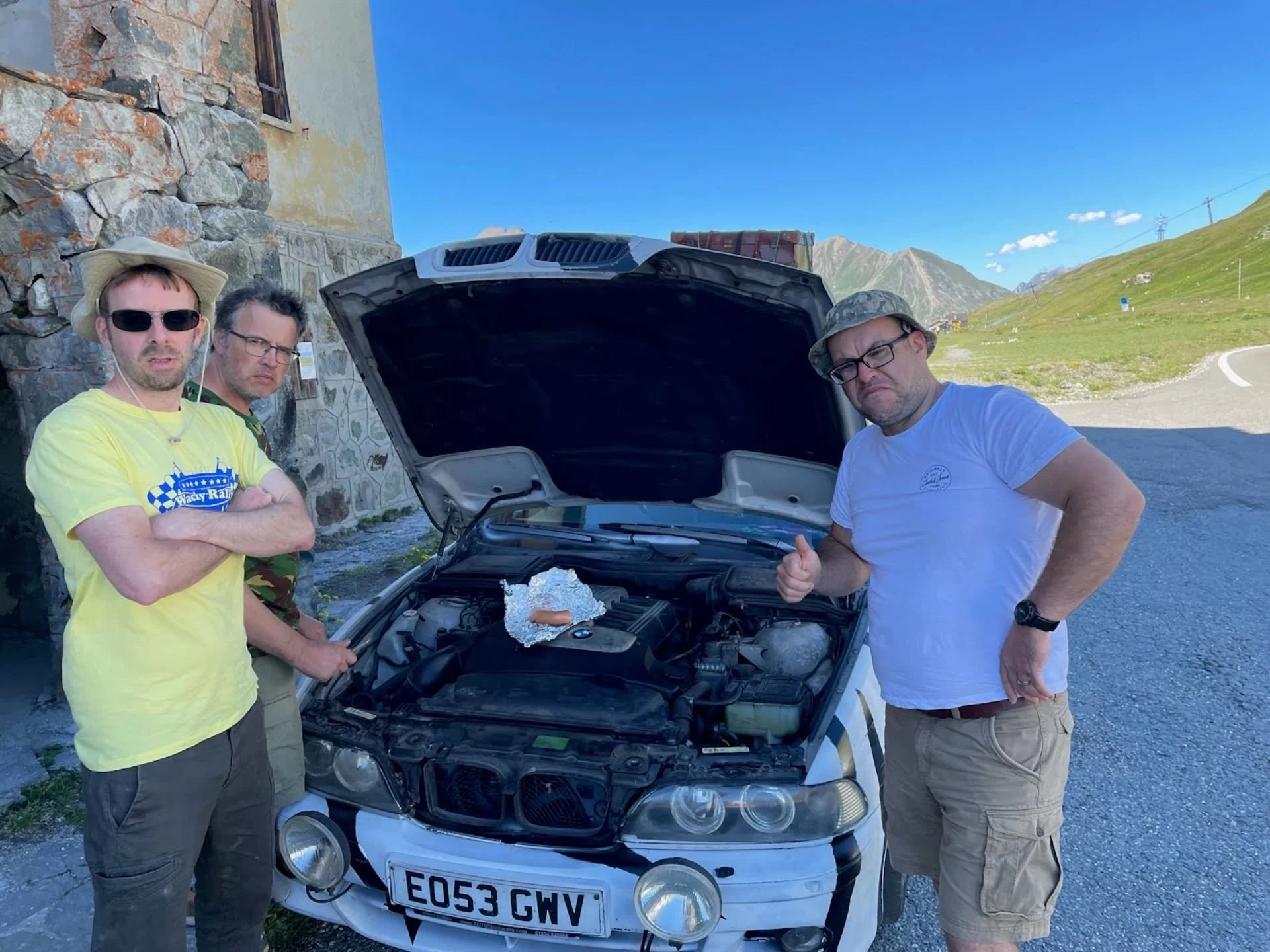 Three men stand next to a car with its hood open, on a mountain road. They are outdoors with mountains and blue sky in the background. One man wears a yellow shirt and sunglasses, another wears glasses and a camouflage shirt, and the third wears a wh
