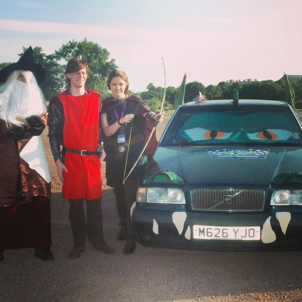 Three people in costume standing next to a black car with eyes and nose painted on the hood, adorned with leaves and a purple flower, on a paved area near water and trees.