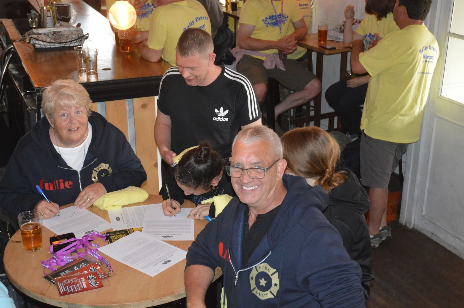 Group of people sitting and standing around a wooden table, signing documents, with some wearing yellow shirts and one wearing a black Adidas shirt, in a casual indoor setting, likely a community or work event.