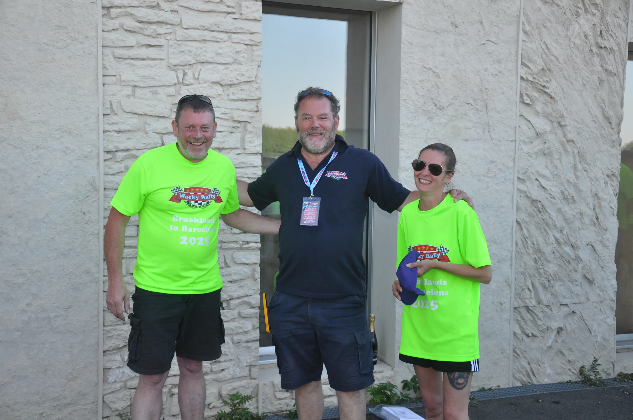 Three smiling people standing outdoors in front of a building wall, with two of them wearing bright yellow T-shirts with event details, and the man in the middle wearing a black polo shirt with a lanyard. They appear to be at a rally or event.