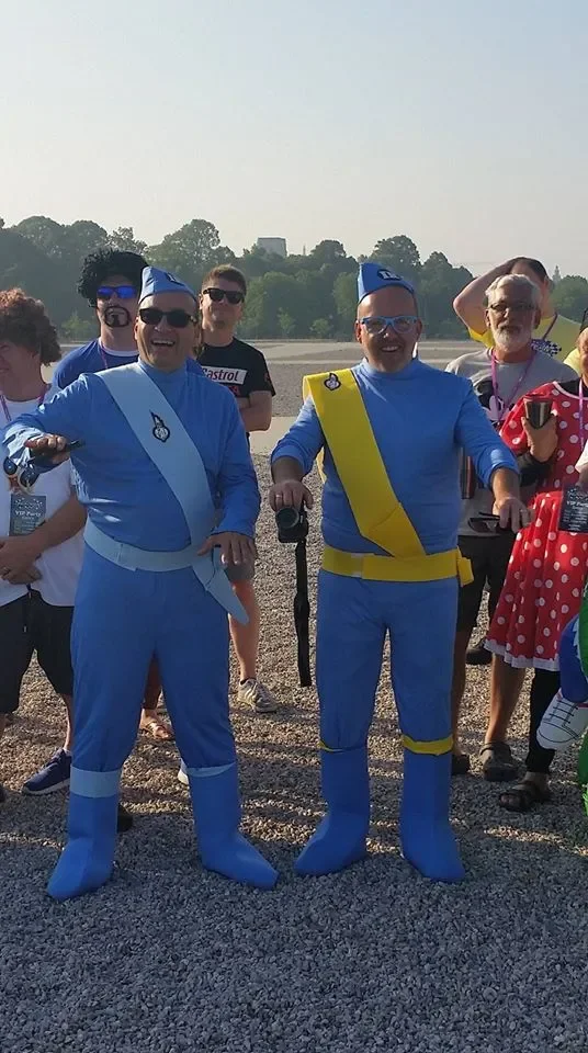 Group of people dressed in costumes, including two men in inflatable blue and yellow outfits resembling cartoon characters, standing outdoors on a gravel surface with trees in the background.