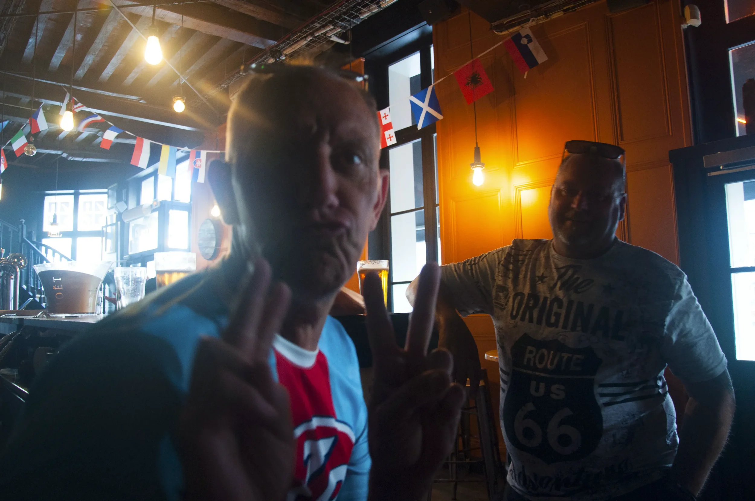 Two men in a pub or bar with flags hanging from the ceiling; one holding up a peace sign and the other wearing sunglasses on his head.