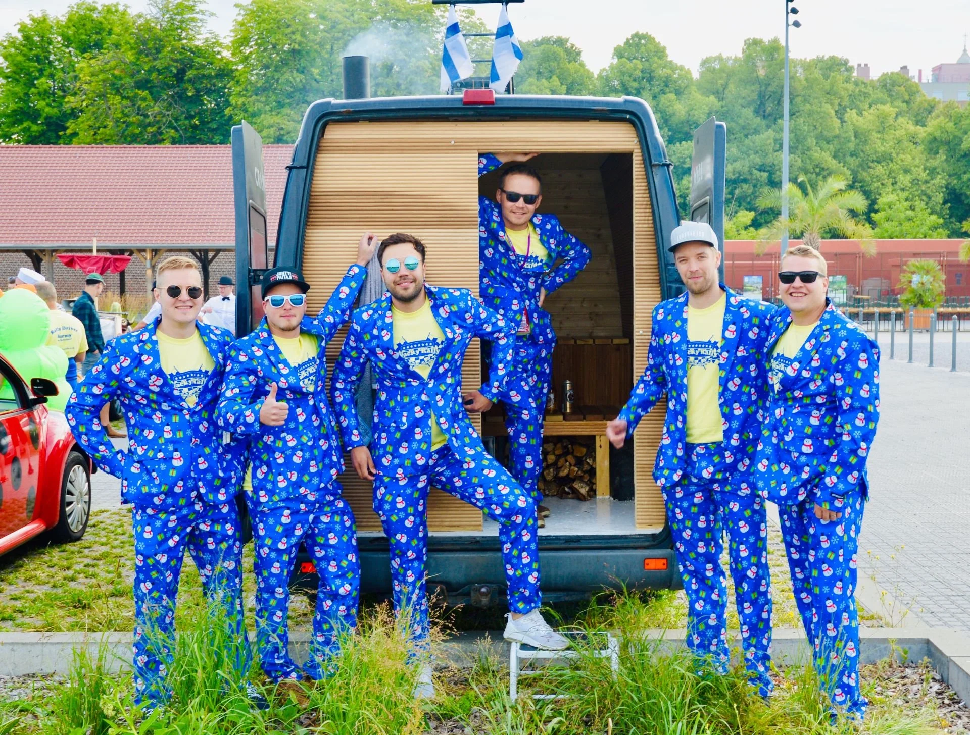 A group of seven men dressed in matching blue pajamas with snowmen and Christmas trees' pattern, standing in front of a food truck with a wooden interior. Some are wearing sunglasses, and one is giving a thumbs-up. The food truck and the men are outd