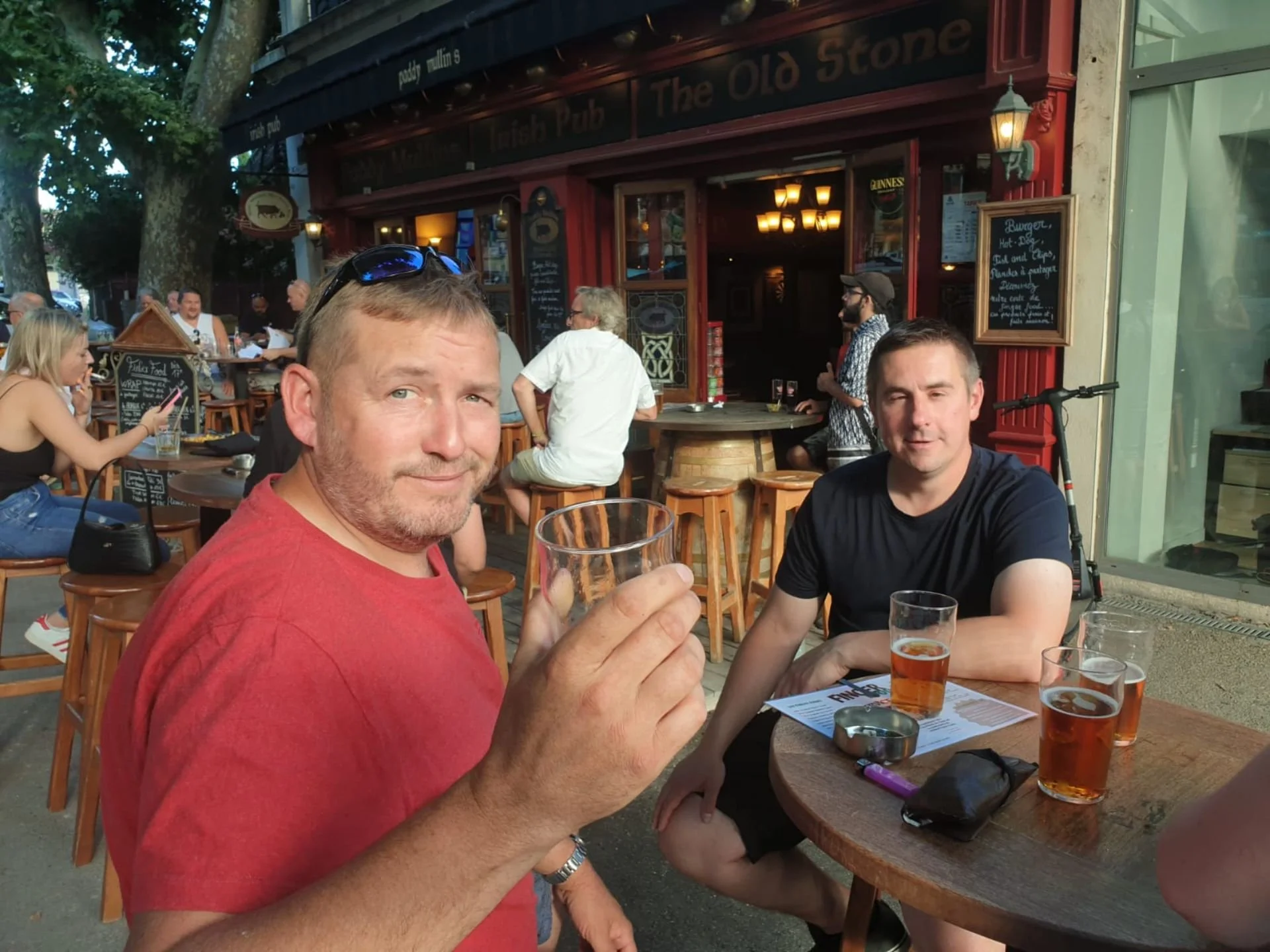 Two men sitting outside a pub, one holding a glass of drink and the other with a drink in front of him, with more patrons seated at tables in the background, in front of a red traditional pub with a black sign.